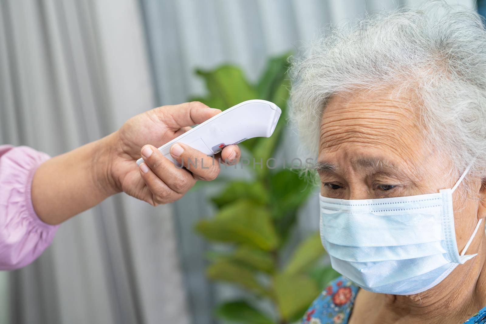 Caregiver holding digital thermometer to measures asian senior or elderly old lady woman patient wearing a face mask have a fever in hospital, healthy strong medical concept.
