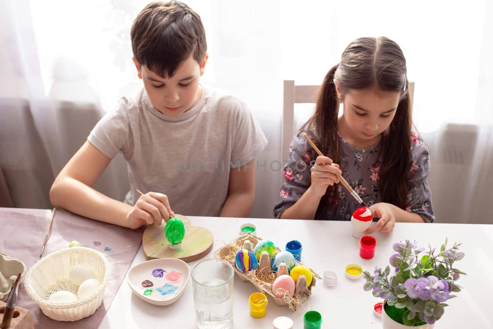 A boy and little girl sits at a white table near the window, prepares for the Easter holiday, paints eggs with a brush.