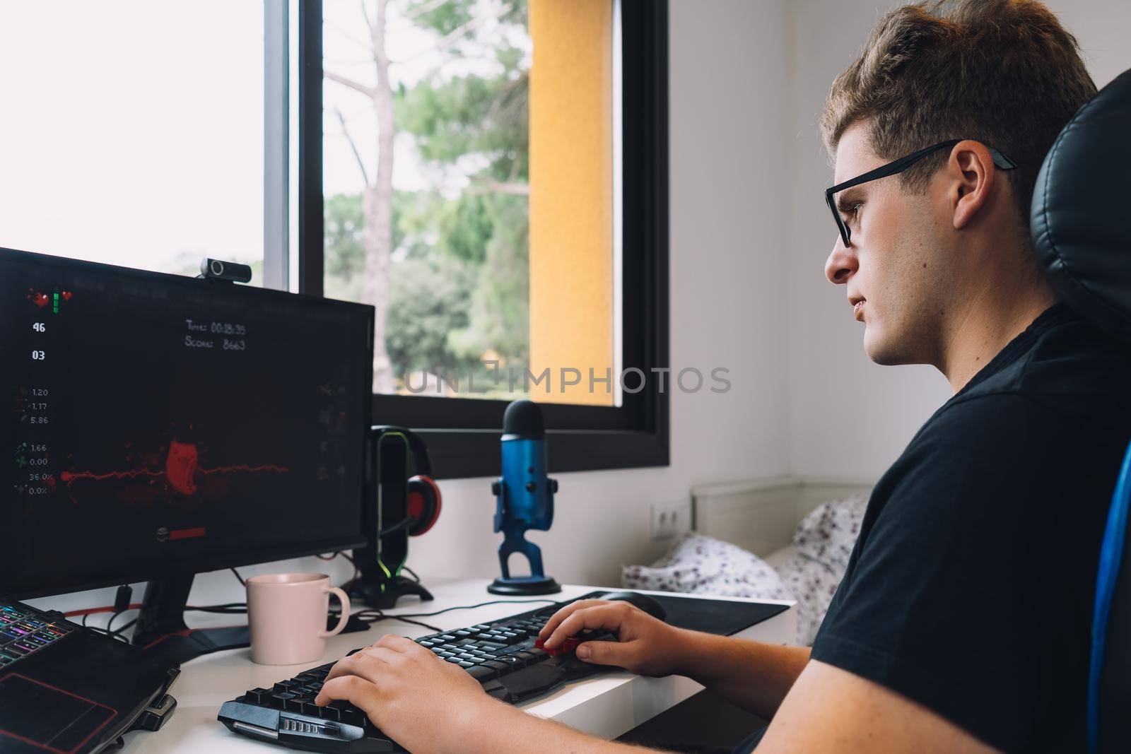 young man with short blond hair wearing glasses, concentrated and hard working, dressed in black shirt, sitting in a gamer chair, working as a graphic designer from home on his computer. natural light from window desk, with computer screen keyboard and coloured lights. Horizontal