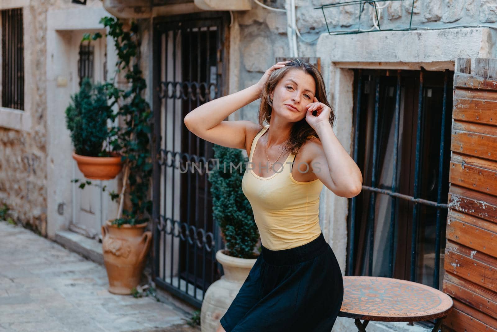 Girl Tourist Walking Through Ancient Narrow Street On A Beautiful Summer Day In MEDITERRANEAN MEDIEVAL CITY, OLD TOWN BUDVA, MONTENEGRO. Young Beautiful Cheerful Woman Walking On Old Street At Tropical Town. Pretty Girl Looking At You And Smiling by Andrii_Ko
