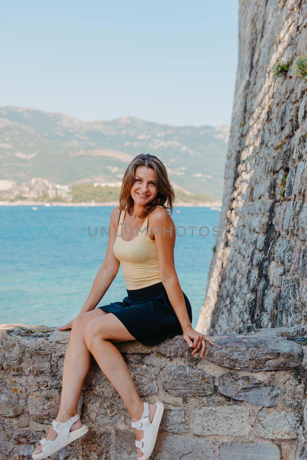 Beautiful girl sitting on a stone wall, in background is the blue sea, Budva, Montenegro. by Andrii_Ko