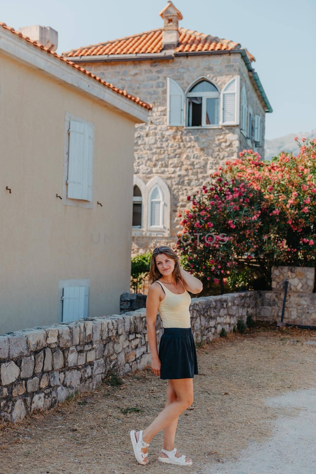 Girl Tourist Walking Through Ancient Narrow Street On A Beautiful Summer Day In MEDITERRANEAN MEDIEVAL CITY, OLD TOWN BUDVA, MONTENEGRO. Young Beautiful Cheerful Woman Walking On Old Street At Tropical Town. Pretty Girl Looking At You And Smiling by Andrii_Ko