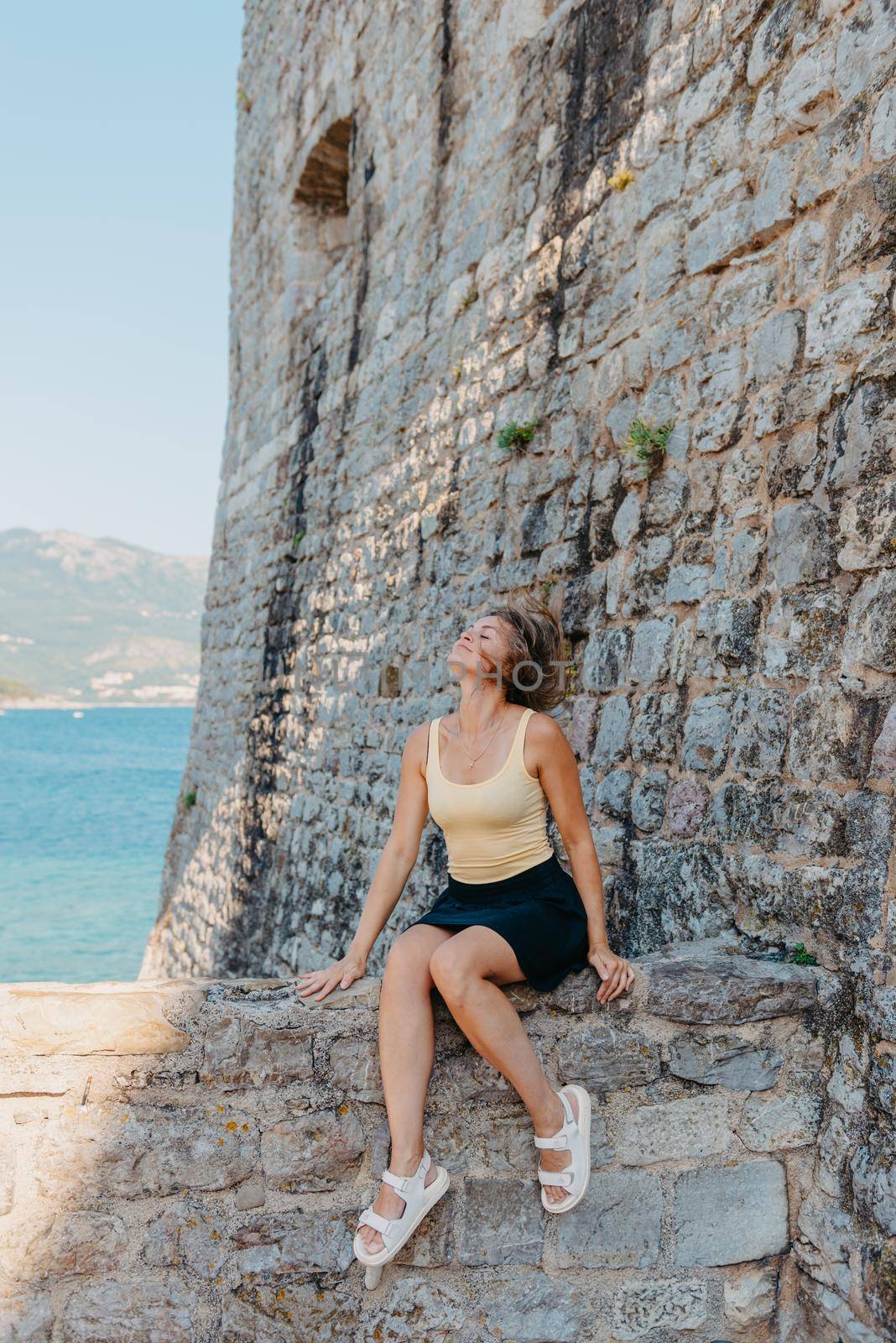 Beautiful girl sitting on a stone wall, in background is the blue sea, Budva, Montenegro