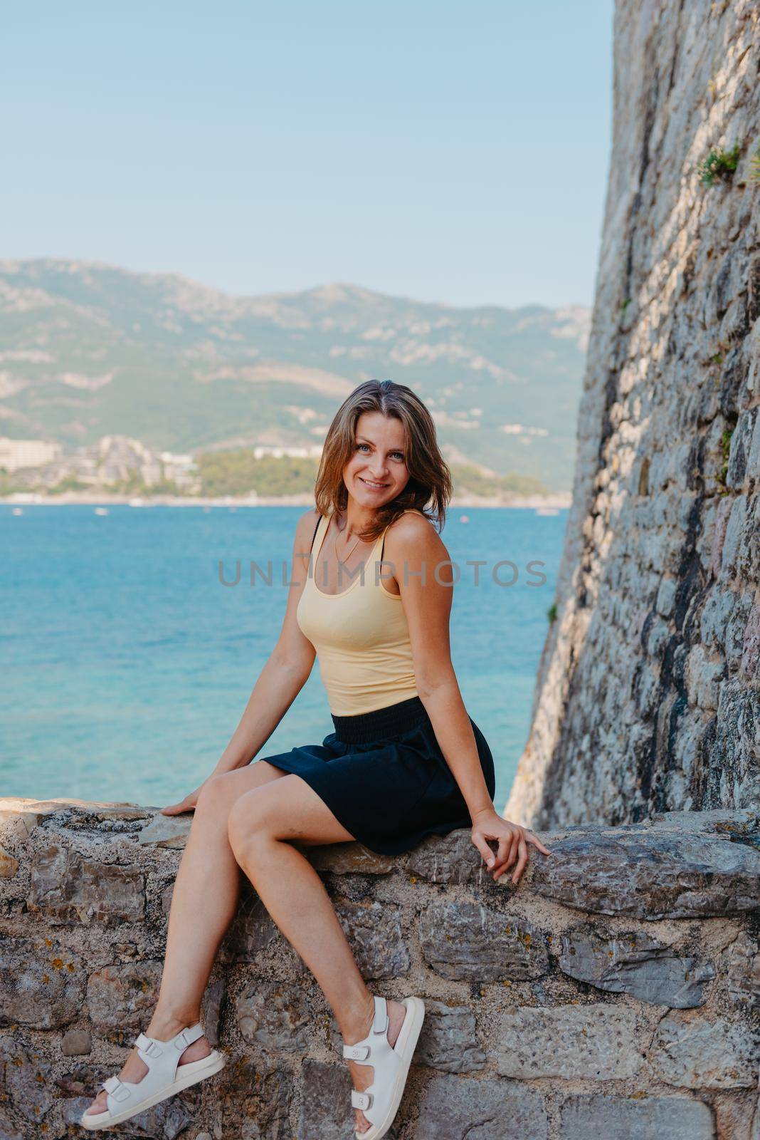 Beautiful girl sitting on a stone wall, in background is the blue sea, Budva, Montenegro. by Andrii_Ko