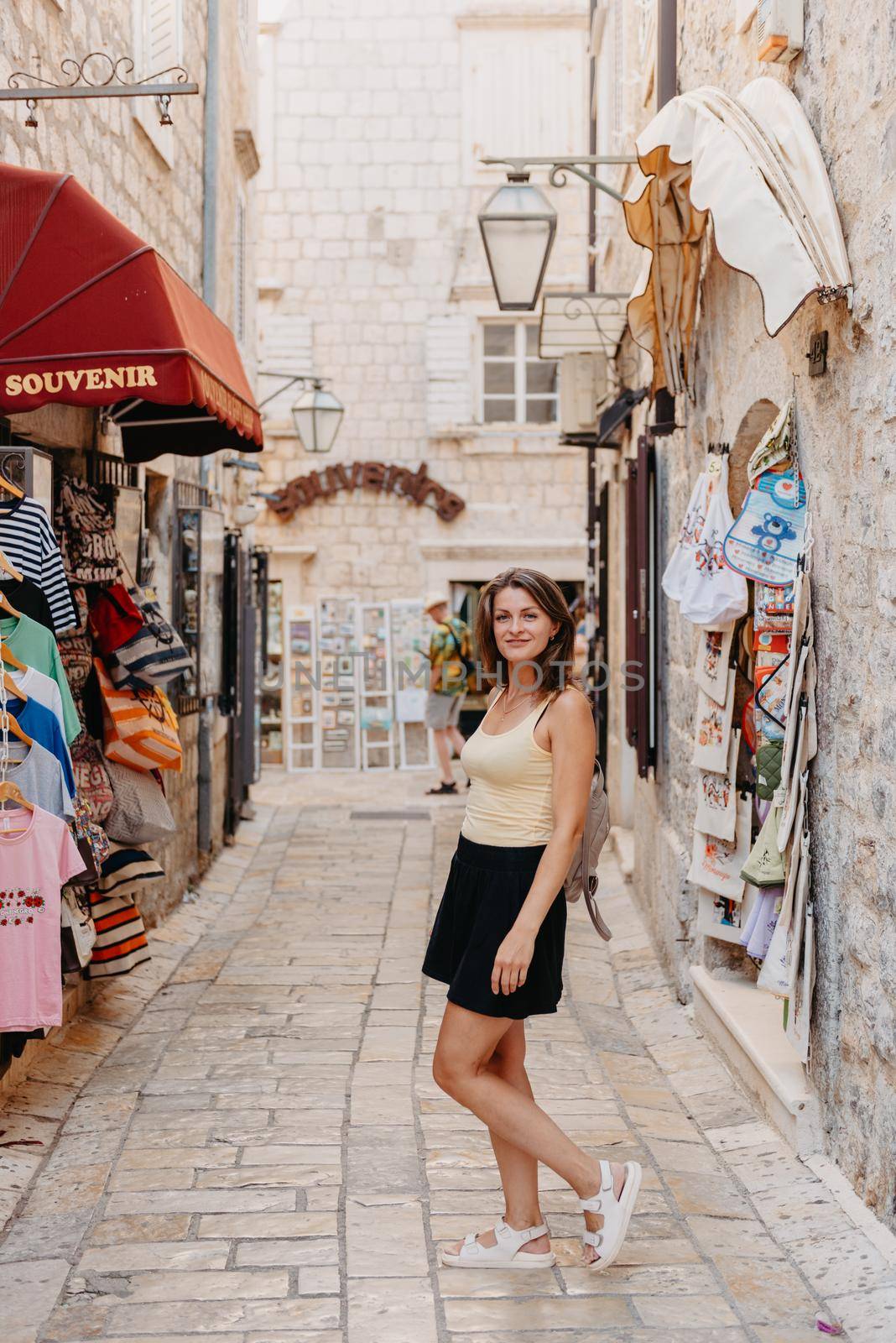 Girl Tourist Walking Through Ancient Narrow Street On A Beautiful Summer Day In MEDITERRANEAN MEDIEVAL CITY, OLD TOWN BUDVA, MONTENEGRO. Young Beautiful Cheerful Woman Walking On Old Street At Tropical Town. Pretty Girl Looking At You And Smiling by Andrii_Ko