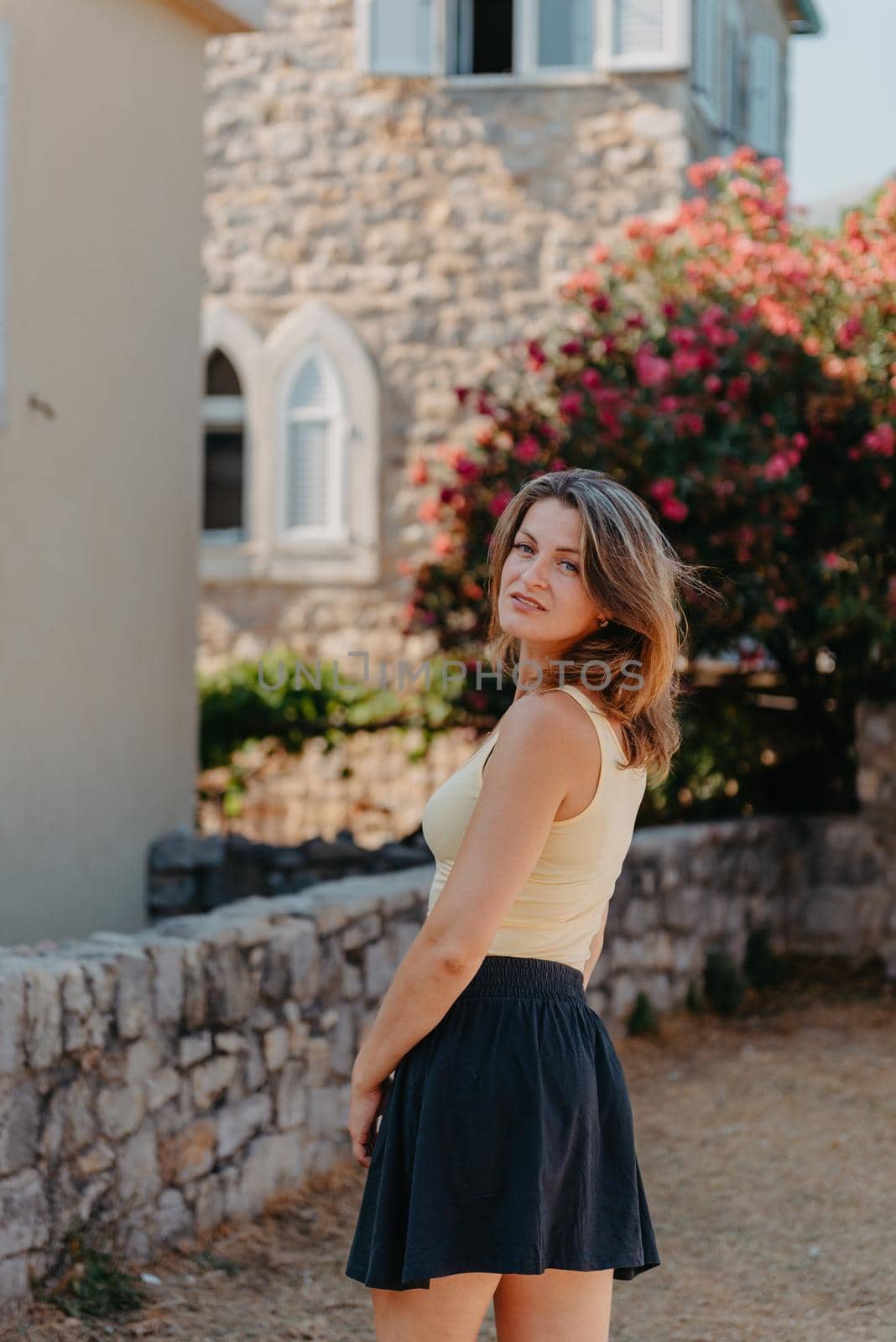 Girl tourist walking through ancient narrow street on a beautiful summer day in MEDITERRANEAN MEDIEVAL CITY , OLD TOWN bUDVA, MONTENEGRO. Young beautiful cheerful woman walking on old street at tropical town. Pretty girl looking at you and smiling