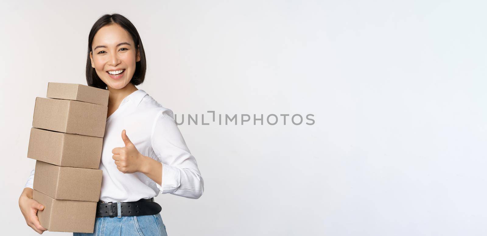 Image of happy modern asian woman showing thumbs up, holding boxes delivery goods, standing against white background.