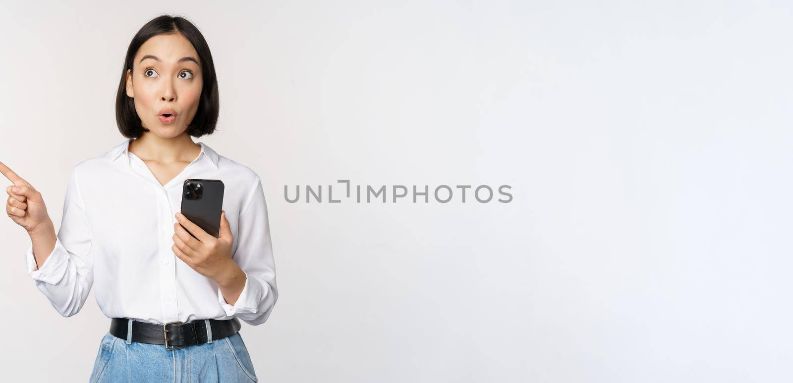 Excited asian girl shopping on mobile app, holding phone and pointing finger left at empty copy space, showing promo text, standing with smartphone against white background.