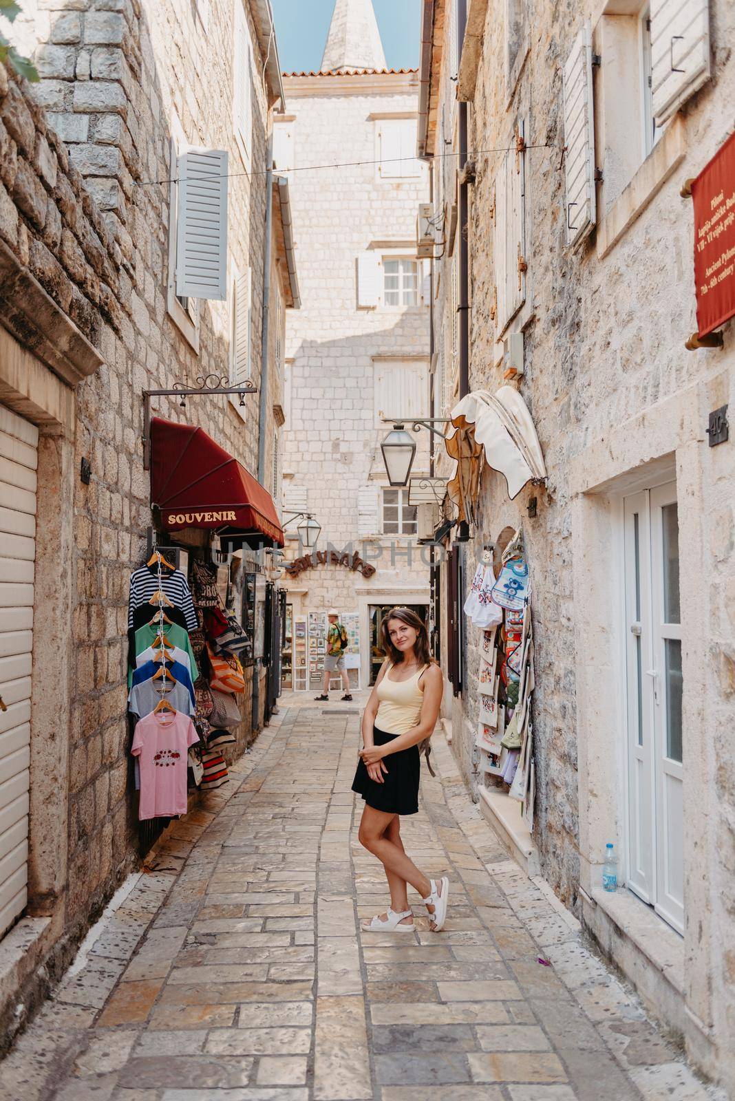 Girl tourist walking through ancient narrow street on a beautiful summer day in MEDITERRANEAN MEDIEVAL CITY , OLD TOWN bUDVA, MONTENEGRO. Young beautiful cheerful woman walking on old street at tropical town. Pretty girl looking at you and smiling