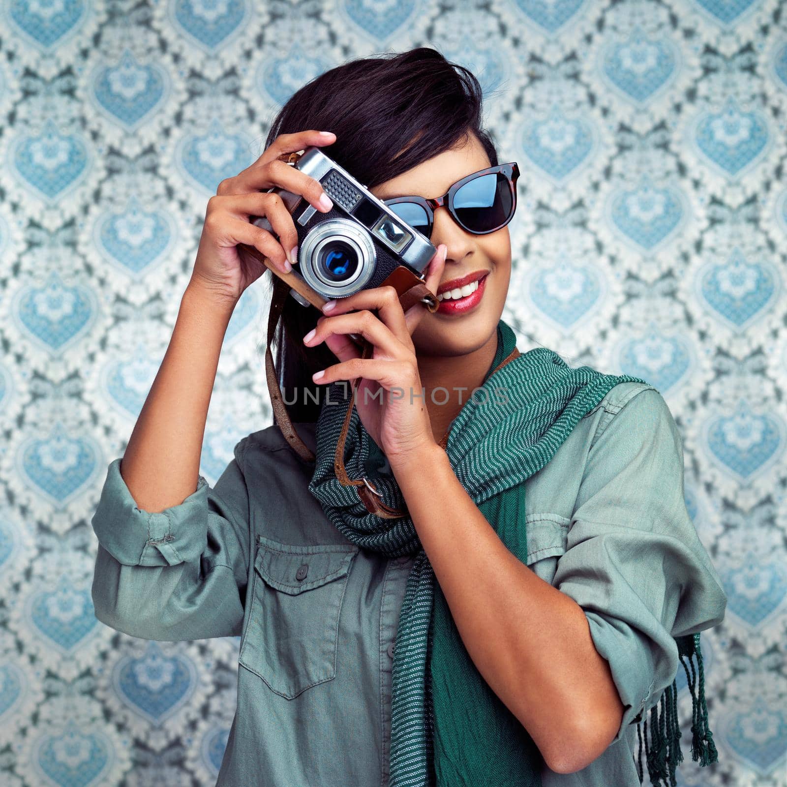 Shot of a young woman posing with a camera over a patterned background.