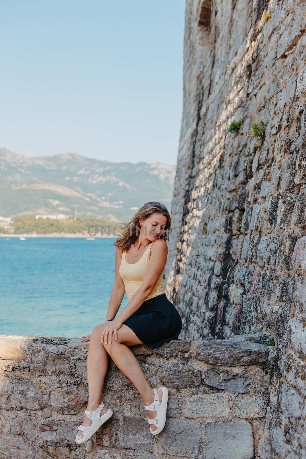 Beautiful girl sitting on a stone wall, in background is the blue sea, Budva, Montenegro