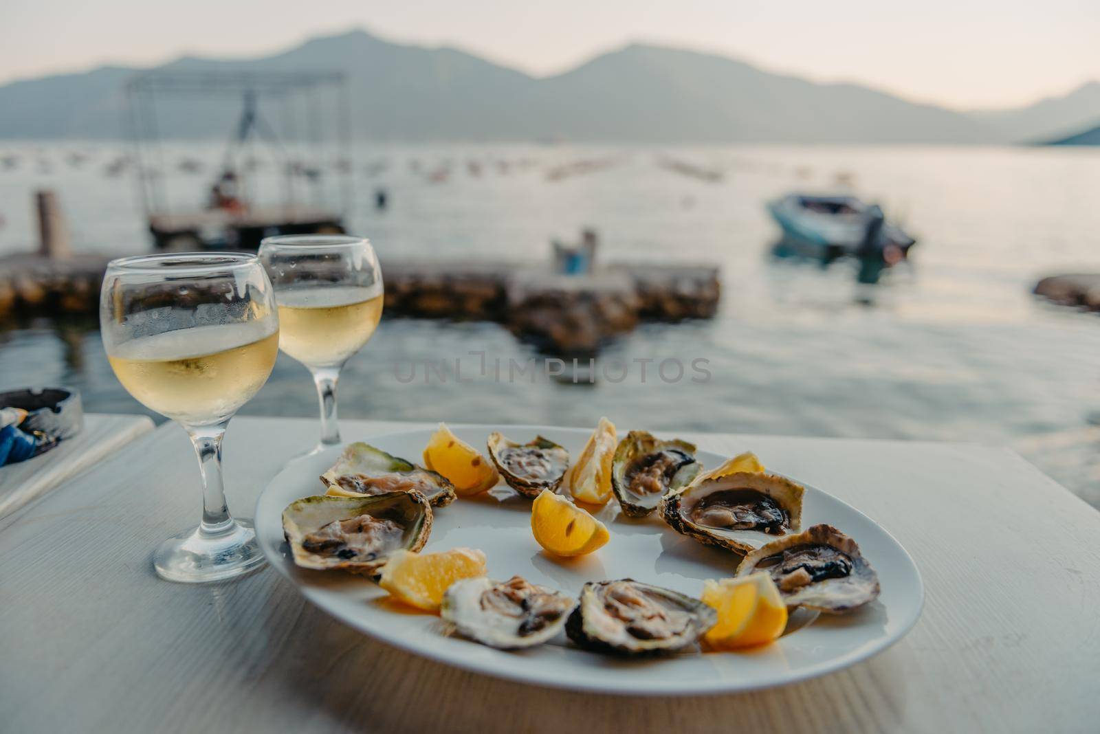 Freshly caught oysters on a plate and vine glasses. Restaurant on the shores of the Bay of Kotor near the oyster farm, Montenegro. Seafood. Beautuful seascape with oyster farm and mountains, Adriatic landscape, Montenegro. Oysters farm in sunset, Boka -Kotor Bay, Montenegro by Andrii_Ko