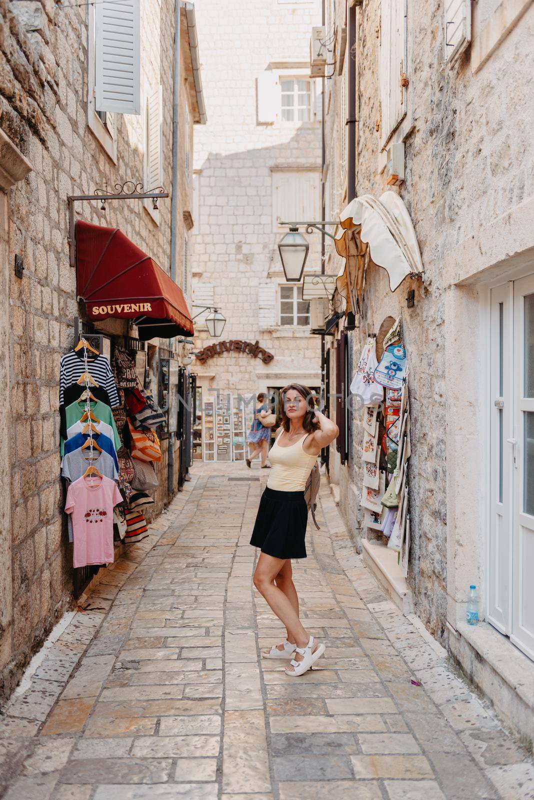 Girl tourist walking through ancient narrow street on a beautiful summer day in MEDITERRANEAN MEDIEVAL CITY , OLD TOWN bUDVA, MONTENEGRO. Young beautiful cheerful woman walking on old street at tropical town. Pretty girl looking at you and smiling