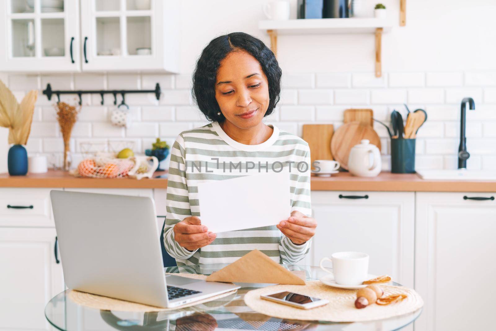 Smiling African-American woman reading paper letter in kitchen. Concept of receiving mail correspondence. Opening envelope. Postage hobby. Looking for news with enthusiasm