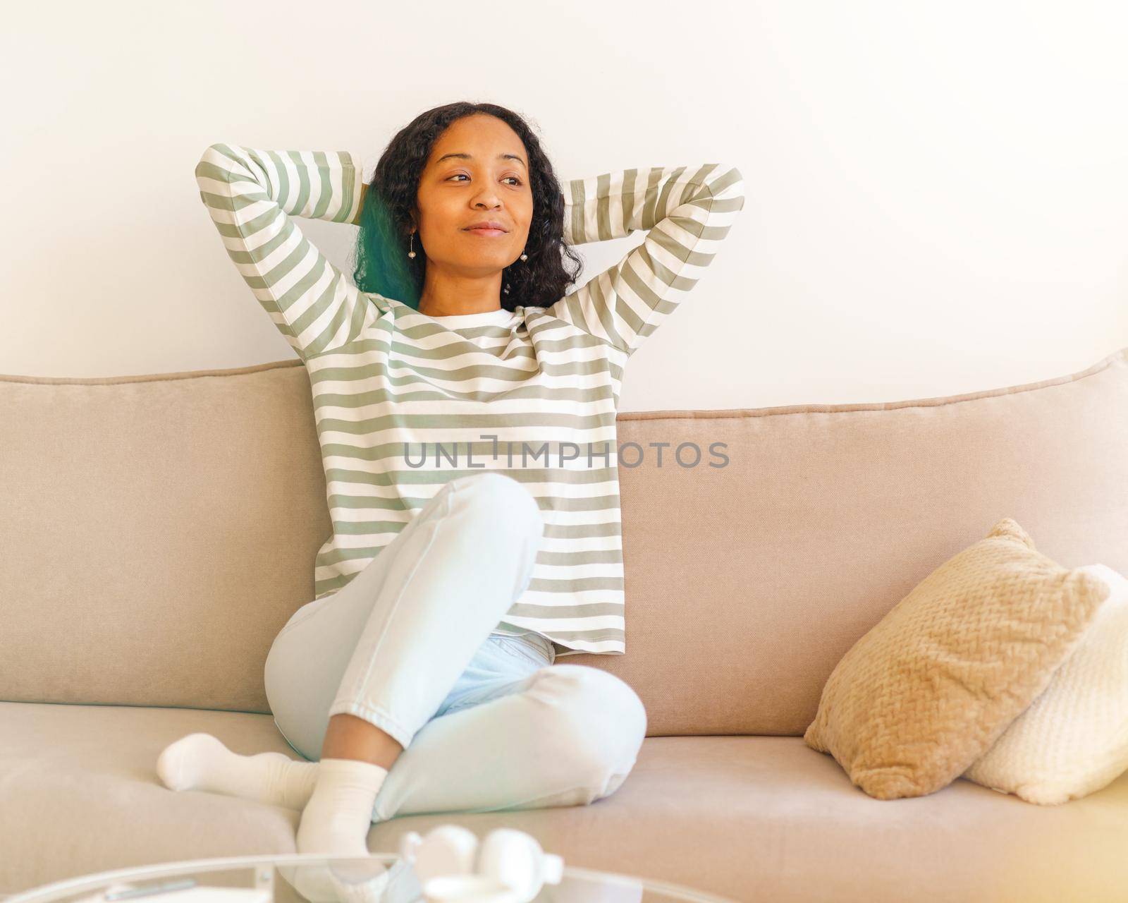 Smiling African-American woman putting hands behind head while sitting on sofa in living room by NataBene