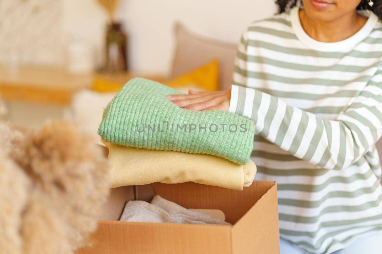 Unrecognizable African-American woman folding clothing in box. Preparing stuff for charity donation. Giving away old things. Sending clothes while relocating. Horizontal, selective focus