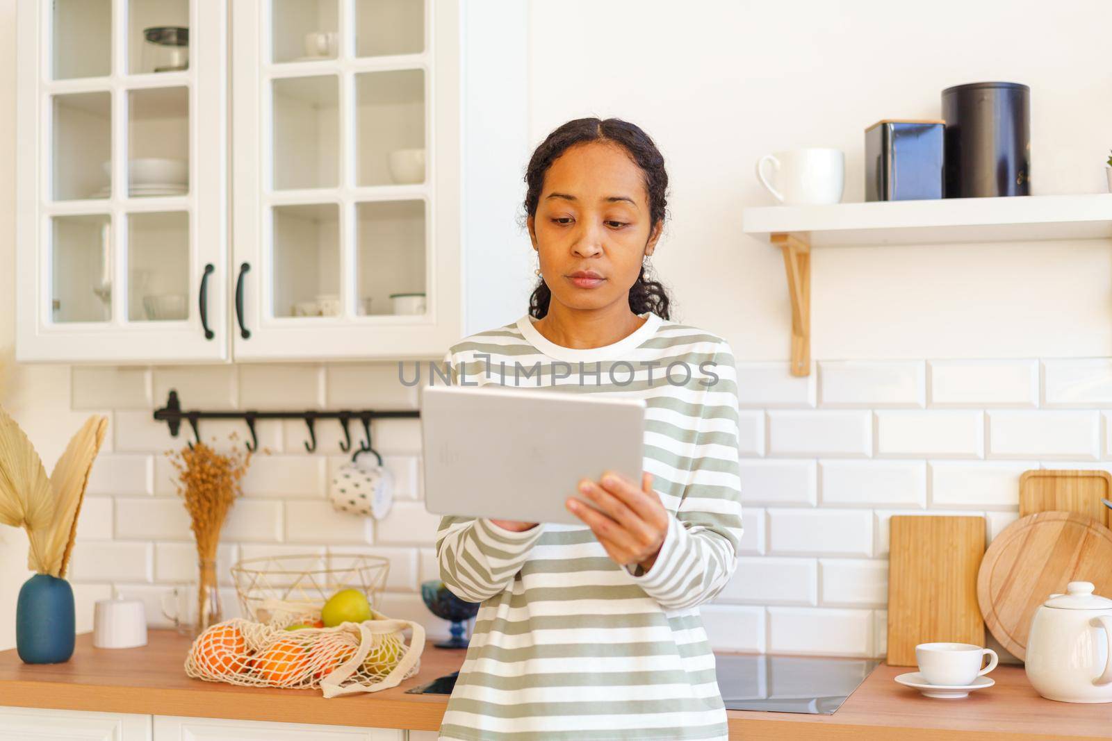 African-American woman using tablet pc while looking for apple recipe before cooking food in kitchen. Concept of modern female watching culinary tutorial on digital device