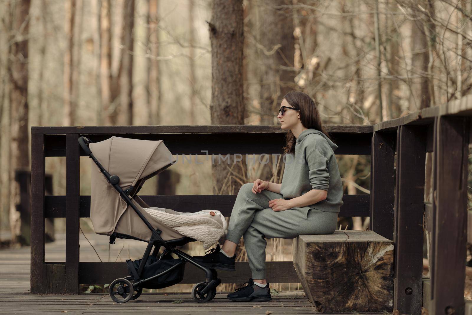 The young mom is relaxing on the wooden bench while her infant child is sleeping in the baby stroller. The mother with her child in the park for fresh air.