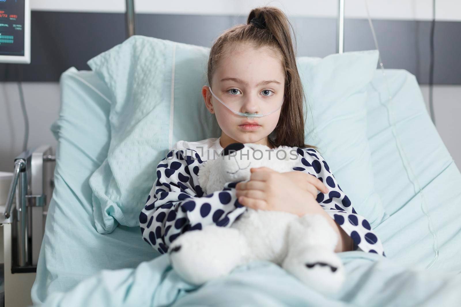 Sick kid holding teddybear and wearing oxygen tube while resting alone in pediatric clinic bed. Unhealthy child in healthcare pediatric hospital patients treatment ward room while looking at camera.