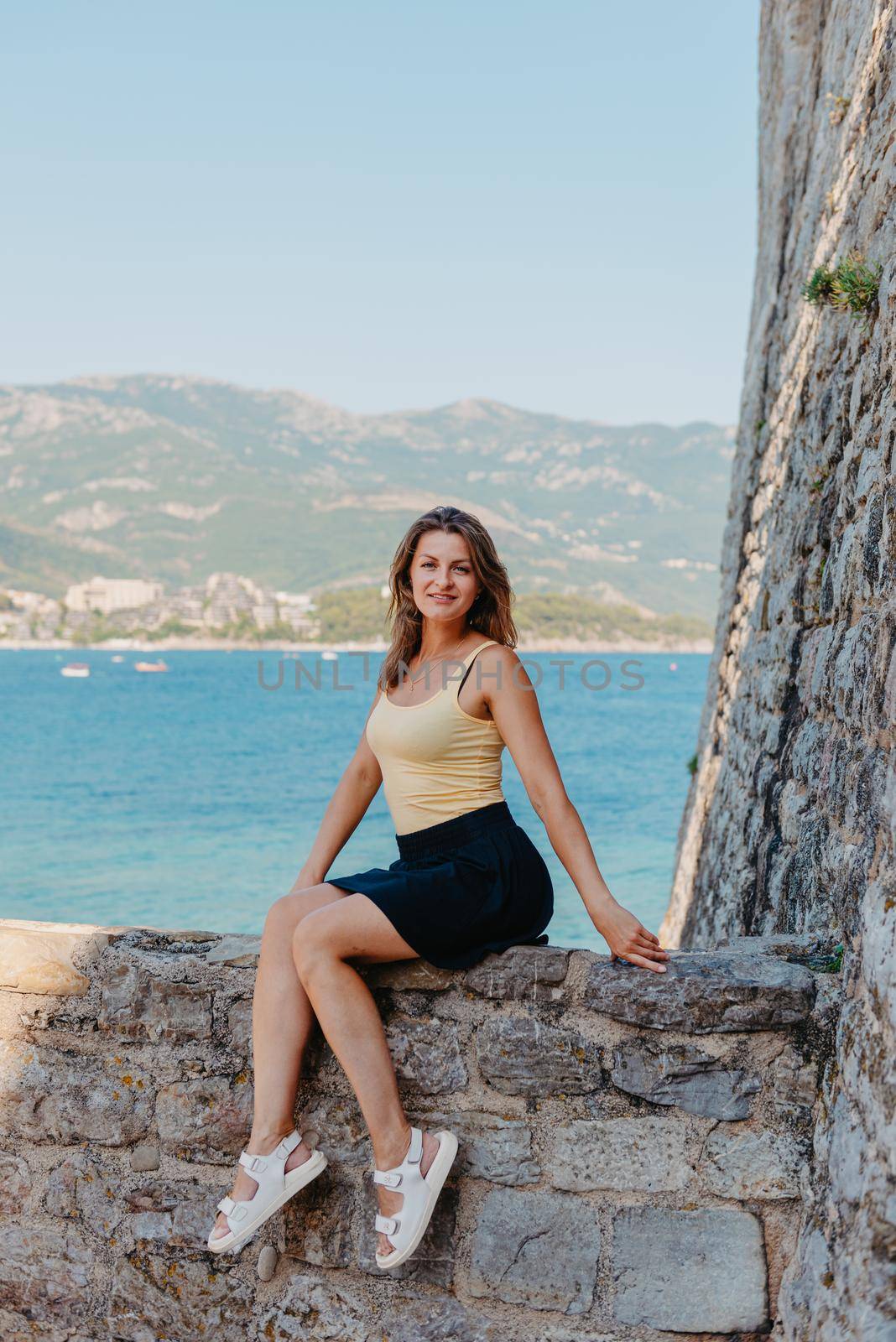 Beautiful girl sitting on a stone wall, in background is the blue sea, Budva, Montenegro