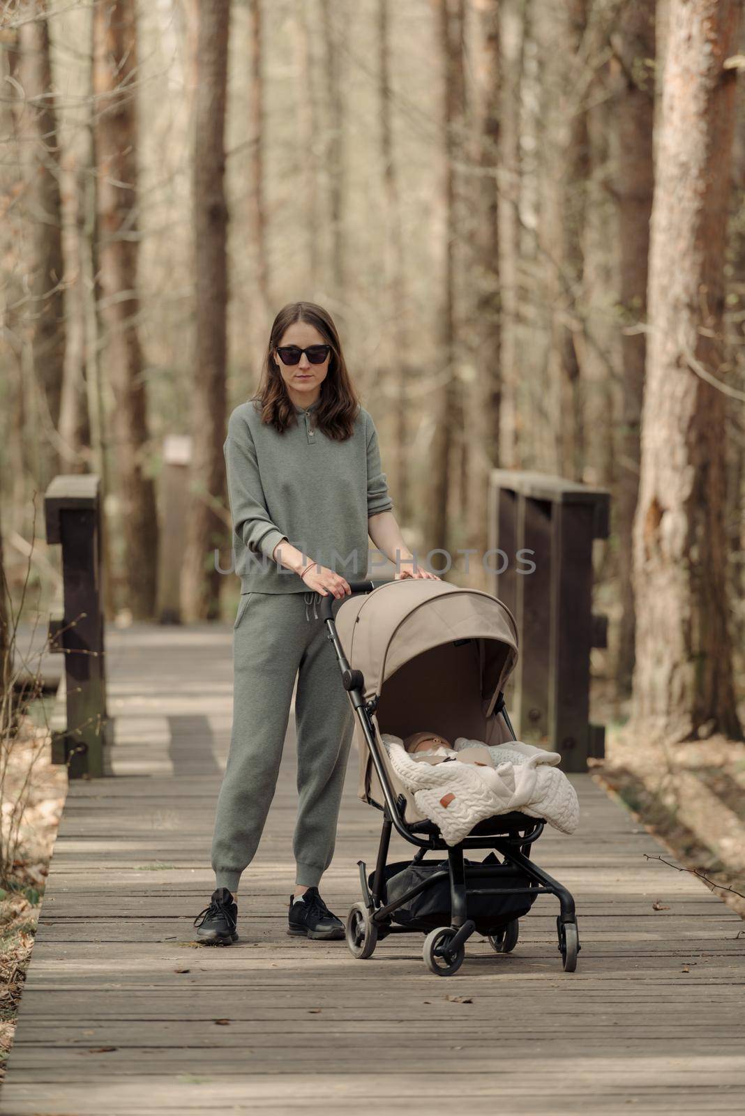 The young mom is walking on the wooden walkway with her infant child who is sleeping in the baby stroller. The mother with her child in the park for fresh air.