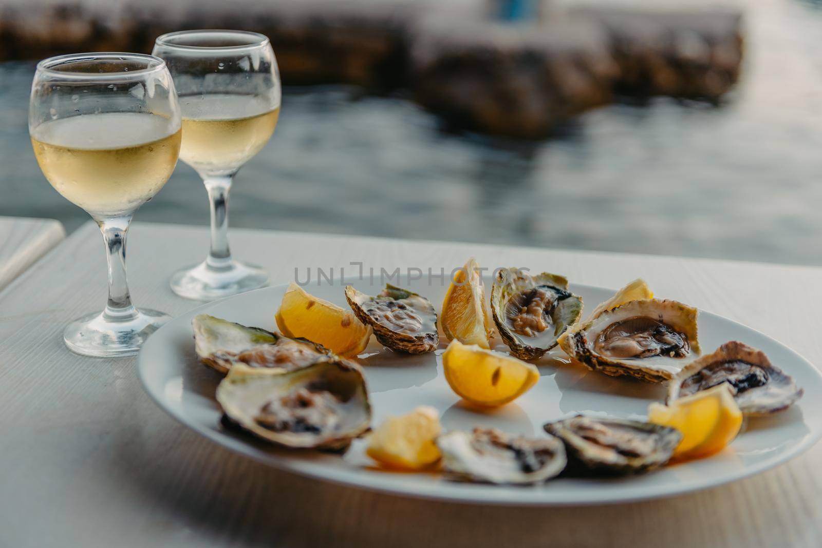 Freshly caught oysters on a plate and vine glasses. Restaurant on the shores of the Bay of Kotor near the oyster farm, Montenegro. Seafood. Beautuful seascape with oyster farm and mountains, Adriatic landscape, Montenegro. Oysters farm in sunset, Boka -Kotor Bay, Montenegro by Andrii_Ko