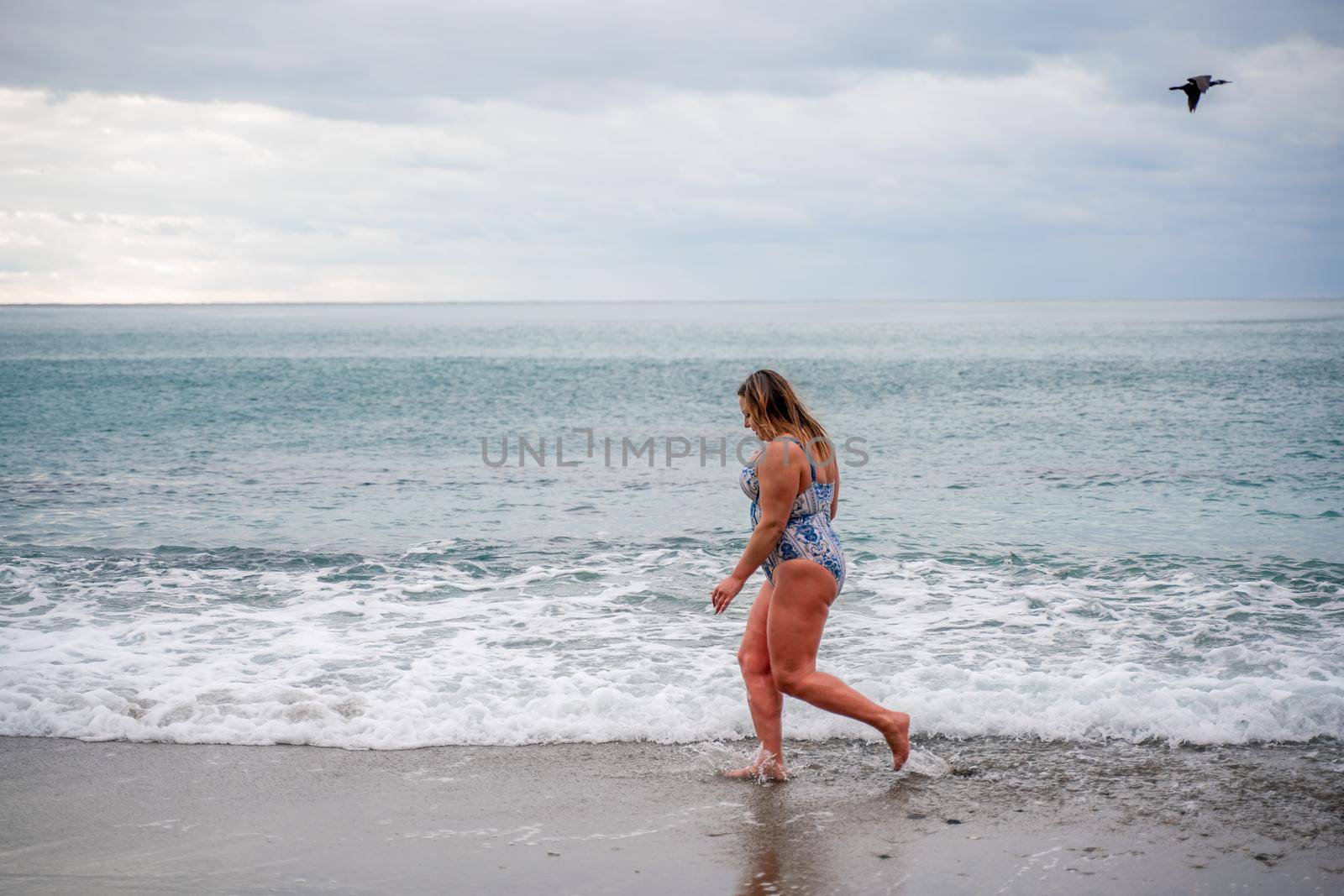 A plump woman in a bathing suit enters the water during the surf. Alone on the beach, Gray sky in the clouds, swimming in winter