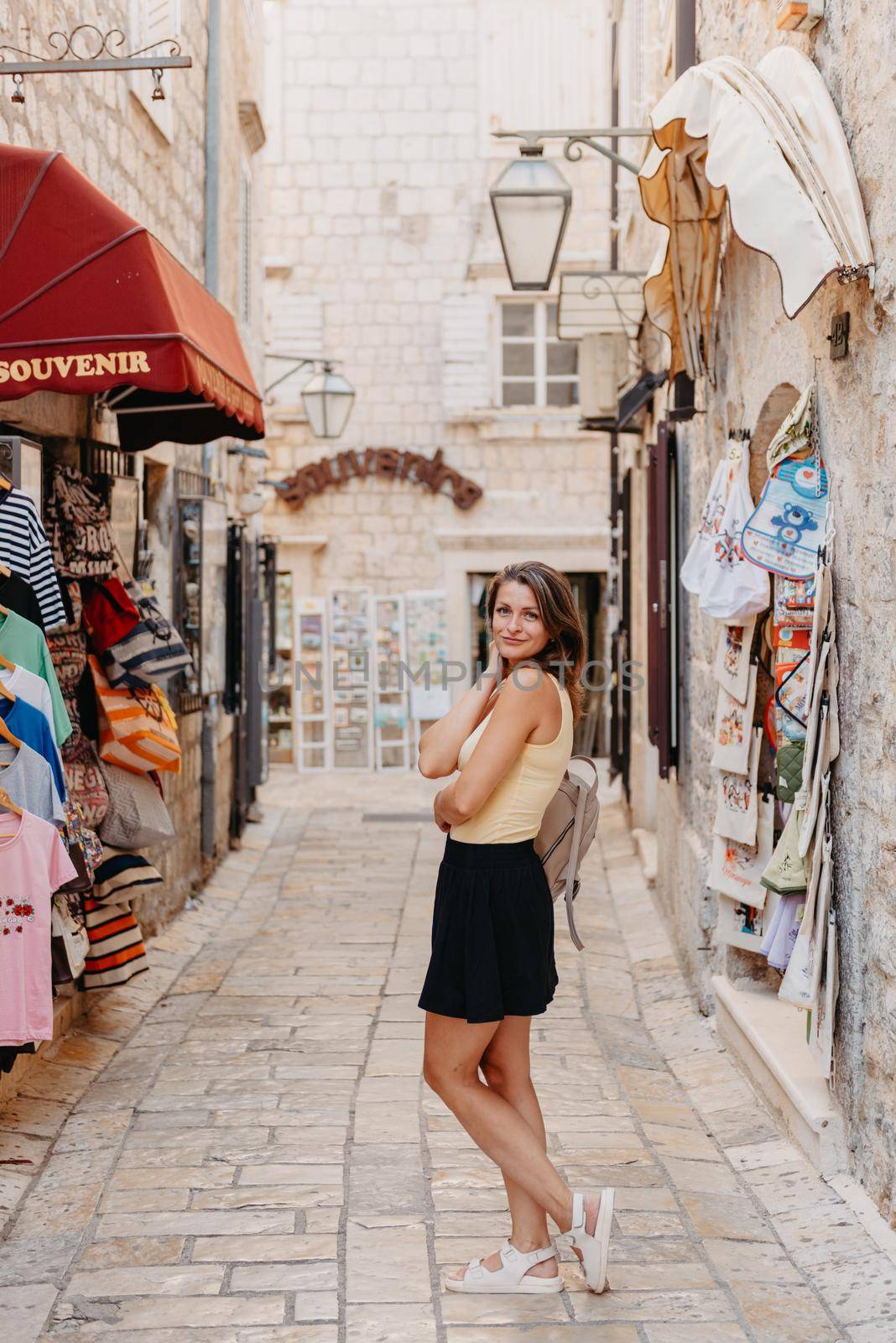 Girl Tourist Walking Through Ancient Narrow Street On A Beautiful Summer Day In MEDITERRANEAN MEDIEVAL CITY, OLD TOWN BUDVA, MONTENEGRO. Young Beautiful Cheerful Woman Walking On Old Street At Tropical Town. Pretty Girl Looking At You And Smiling by Andrii_Ko