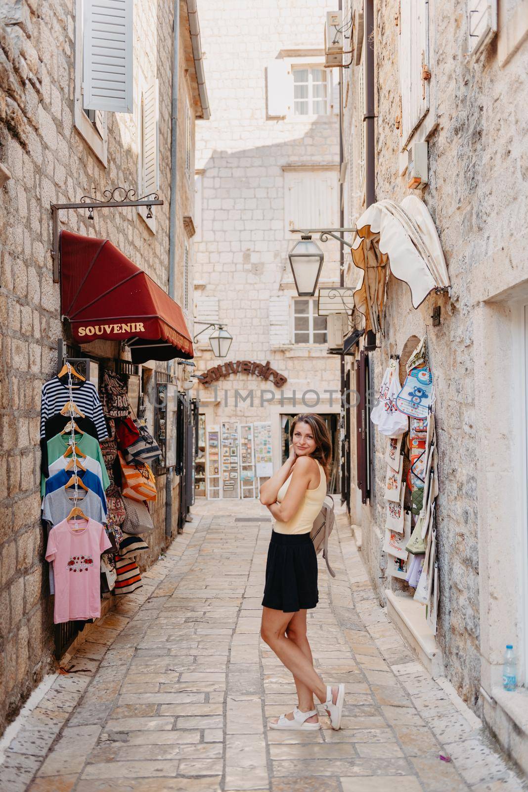 Girl tourist walking through ancient narrow street on a beautiful summer day in MEDITERRANEAN MEDIEVAL CITY , OLD TOWN bUDVA, MONTENEGRO. Young beautiful cheerful woman walking on old street at tropical town. Pretty girl looking at you and smiling