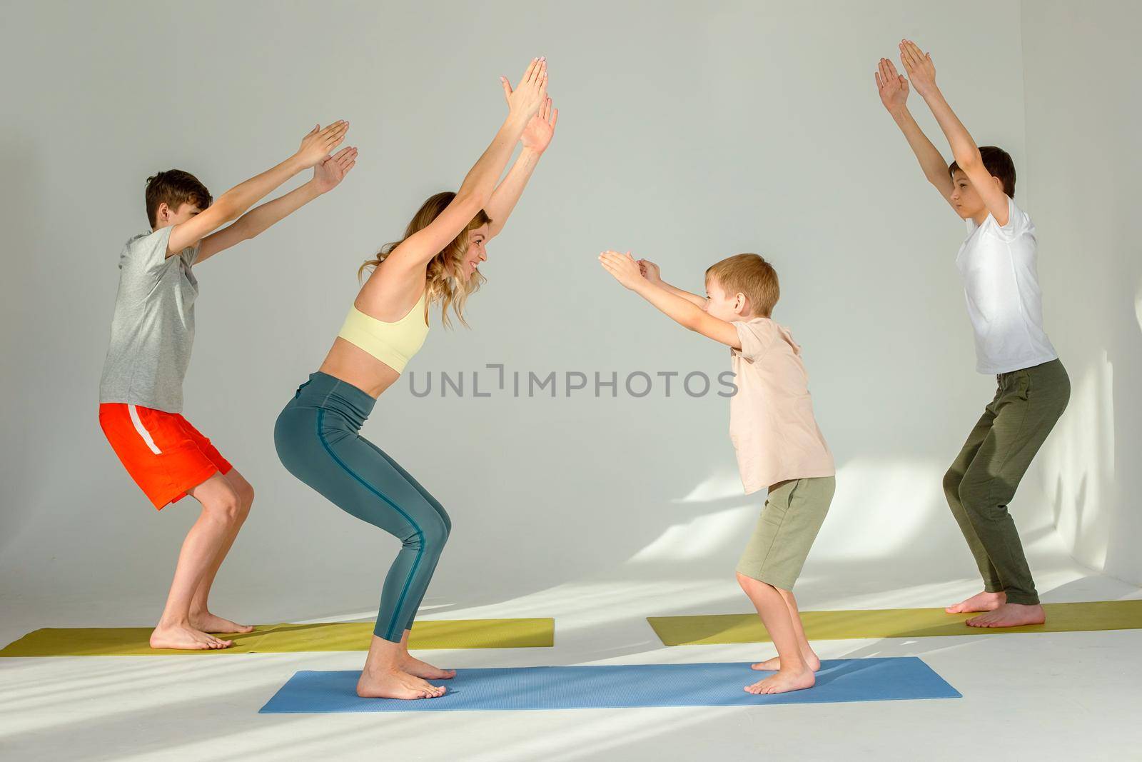 Slim woman, boy and two teenagers perform yoga exercise, stand on mats in chair pose, in sunny studio.