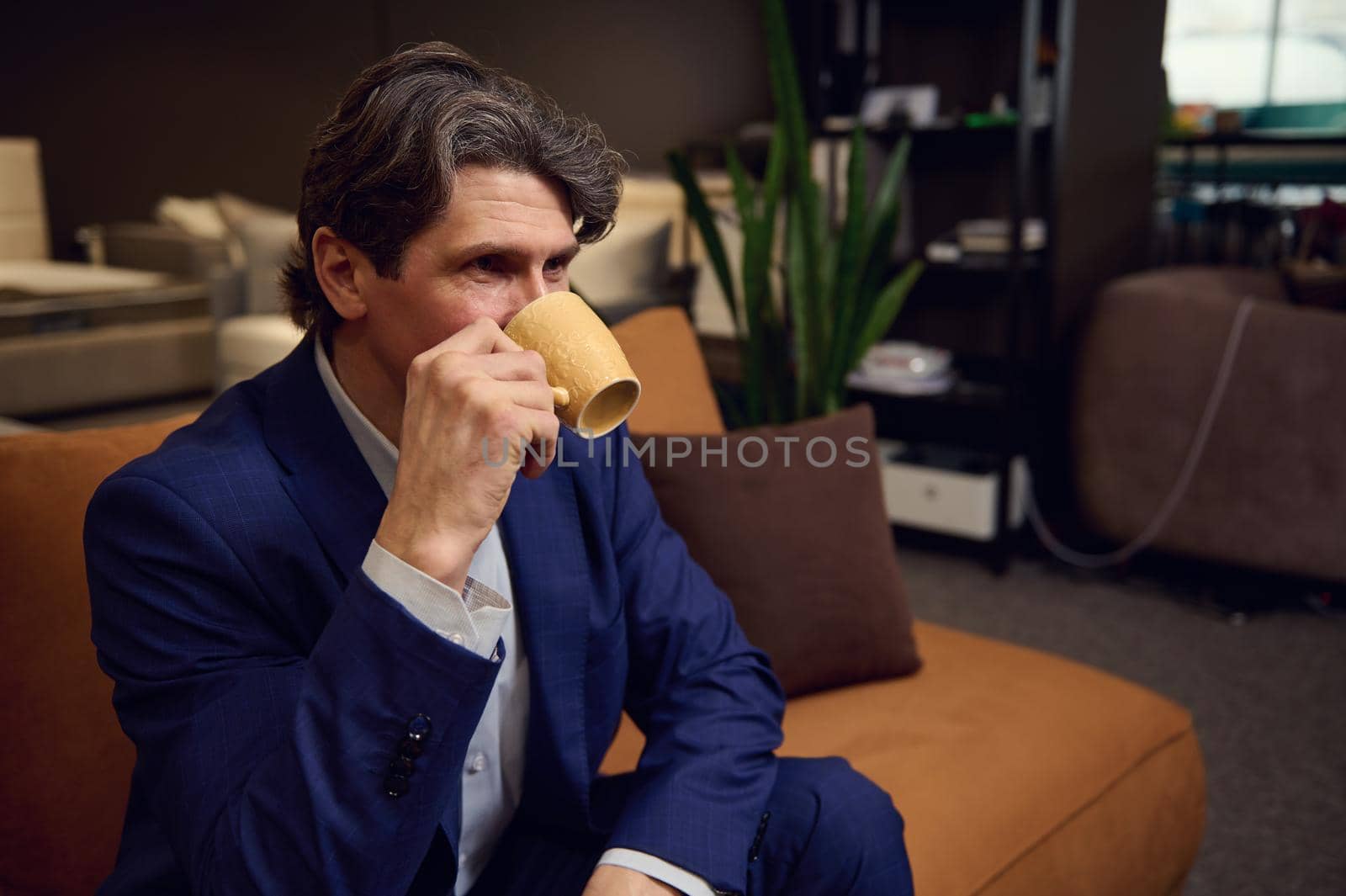 Mature man in business suit drinking coffee, sitting on a sofa in the furniture store showroom. Interior design, house remodelling, home improvement, creative occupation and small business concept