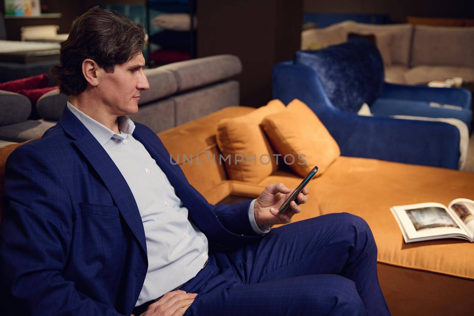 Confident mature man holding mobile phone, reading messages, swiping and surfing on internet websites while sitting on a comfortable sofa with opened catalog of upholstered furniture by artgf