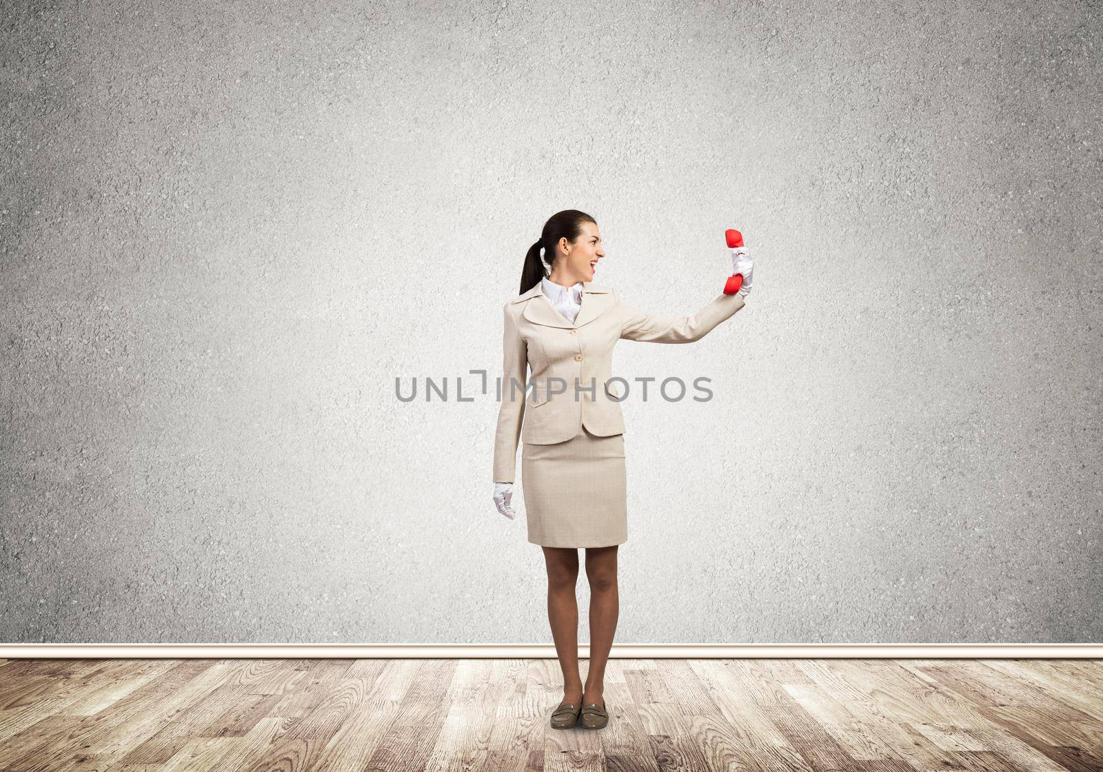 Young angry woman screaming in retro red phone. Emotional call center operator in white business suit posing with phone in room. Hotline support service. Business assistance and consultation.