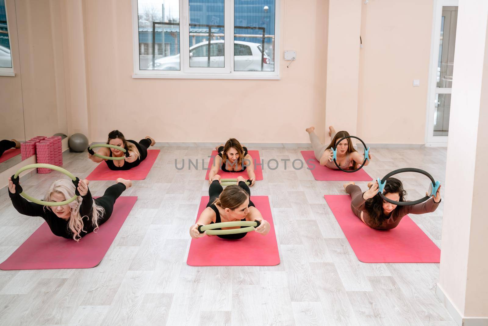 A group of six athletic women doing pilates or yoga on pink mats in front of a window in a beige loft studio interior. Teamwork, good mood and healthy lifestyle concept