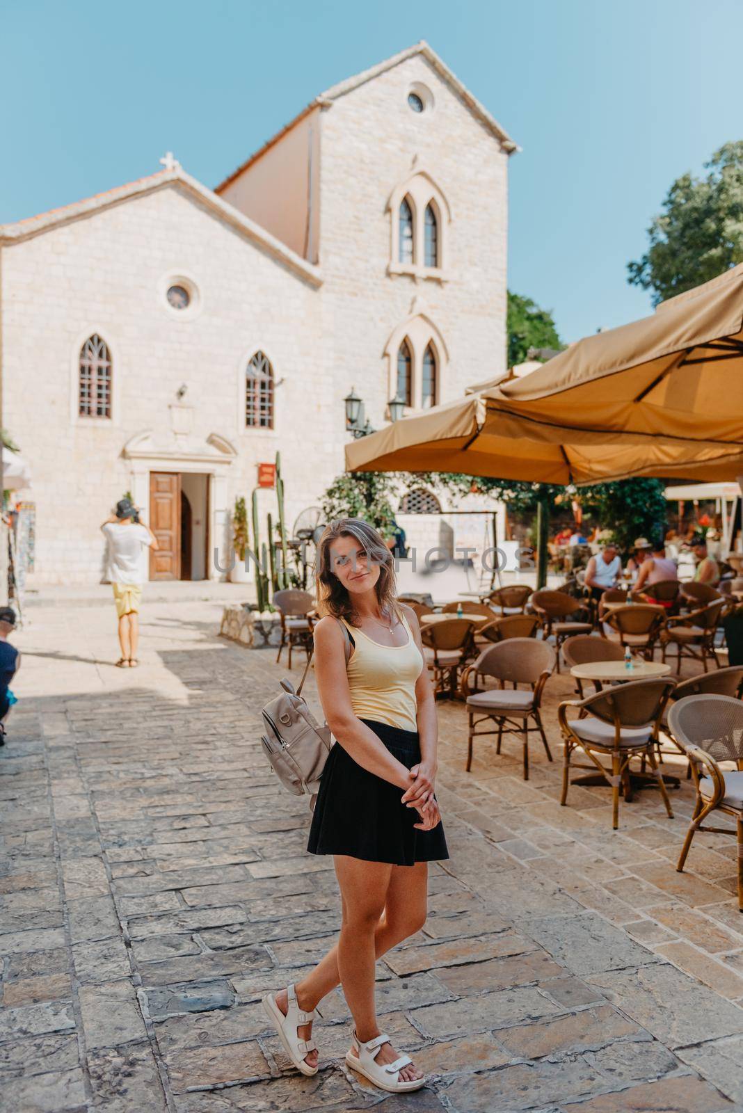 Girl tourist walking through ancient narrow street on a beautiful summer day in MEDITERRANEAN MEDIEVAL CITY , OLD TOWN bUDVA, MONTENEGRO. Young beautiful cheerful woman walking on old street at tropical town. Pretty girl looking at you and smiling