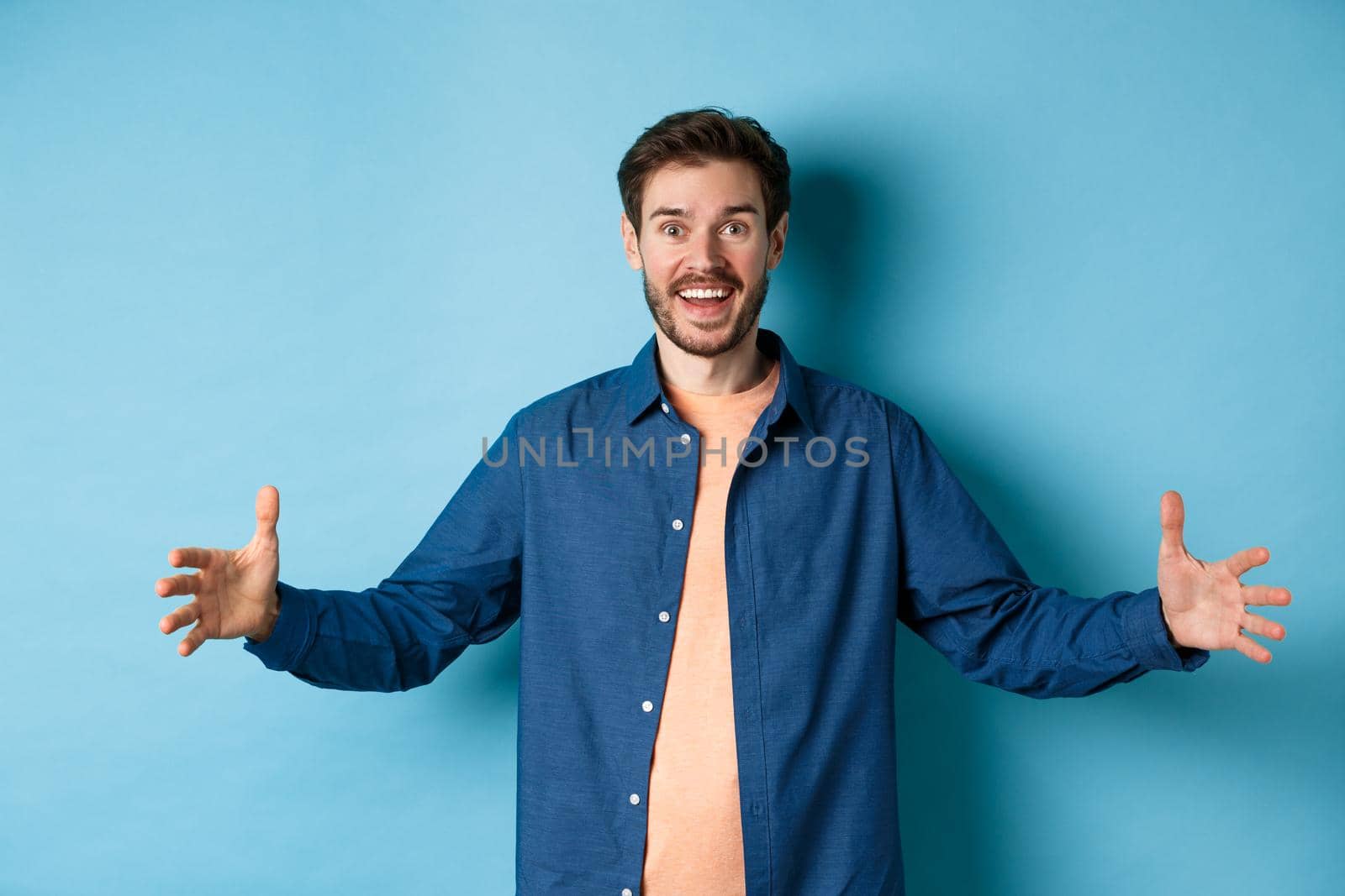 Happy smiling man spread hands sideways and showing big size object, talking about large object, standing cheerful on blue background.