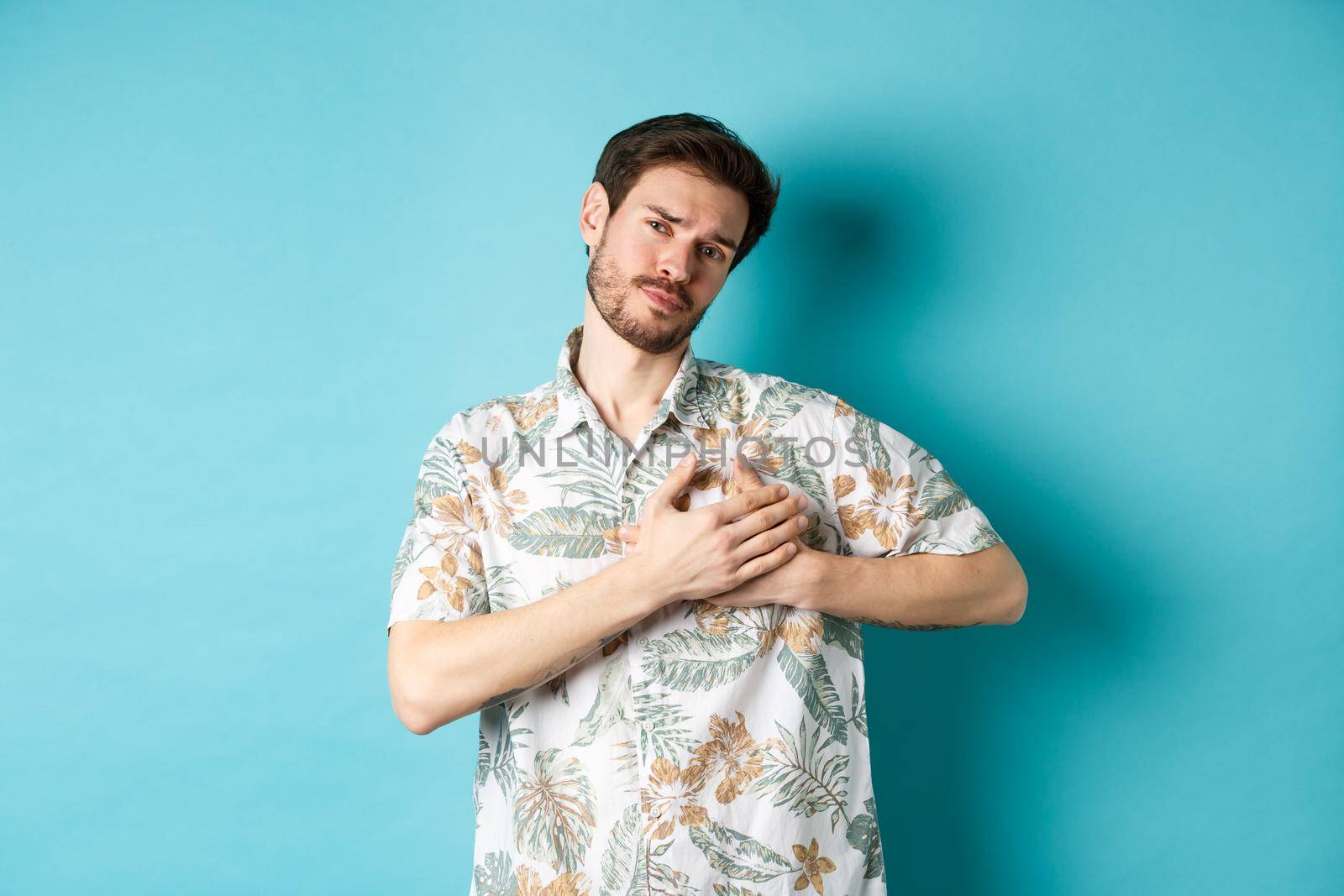 Passionate and romantic guy in hawaiian shirt, holding hands on heart and looking with sympathy, being in love, standing on blue background by Benzoix