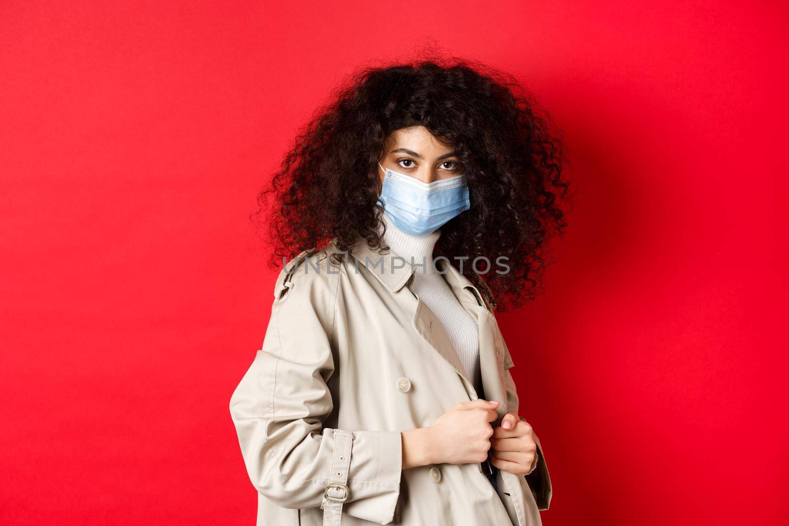 Covid-19, pandemic and quarantine concept. Sassy girl in trench coat and medical mask, put on trench coat for spring walk, red background by Benzoix