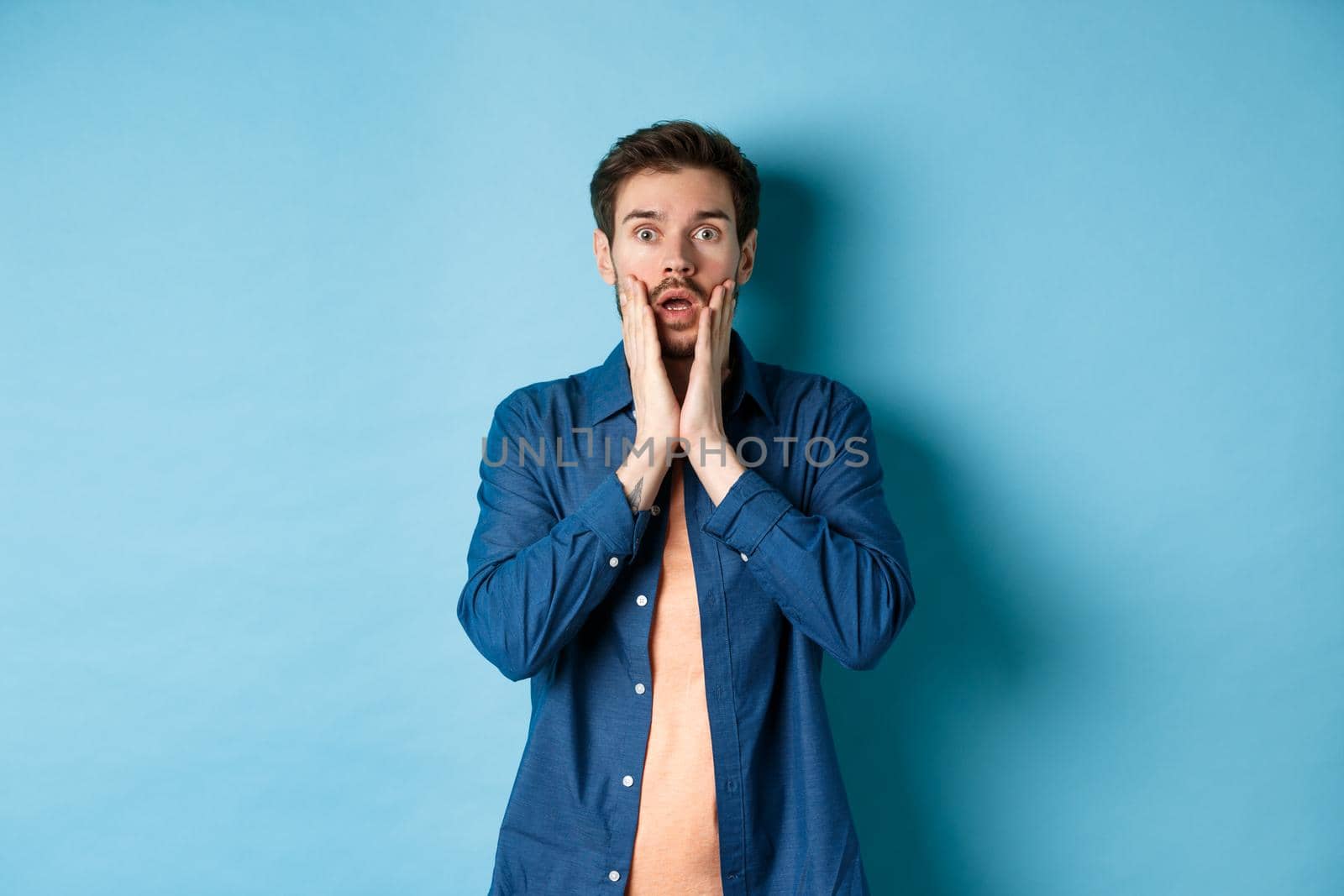 Portrait of guy gasping and dropping jaw with shocked face, staring at camera startled, standing on blue background by Benzoix