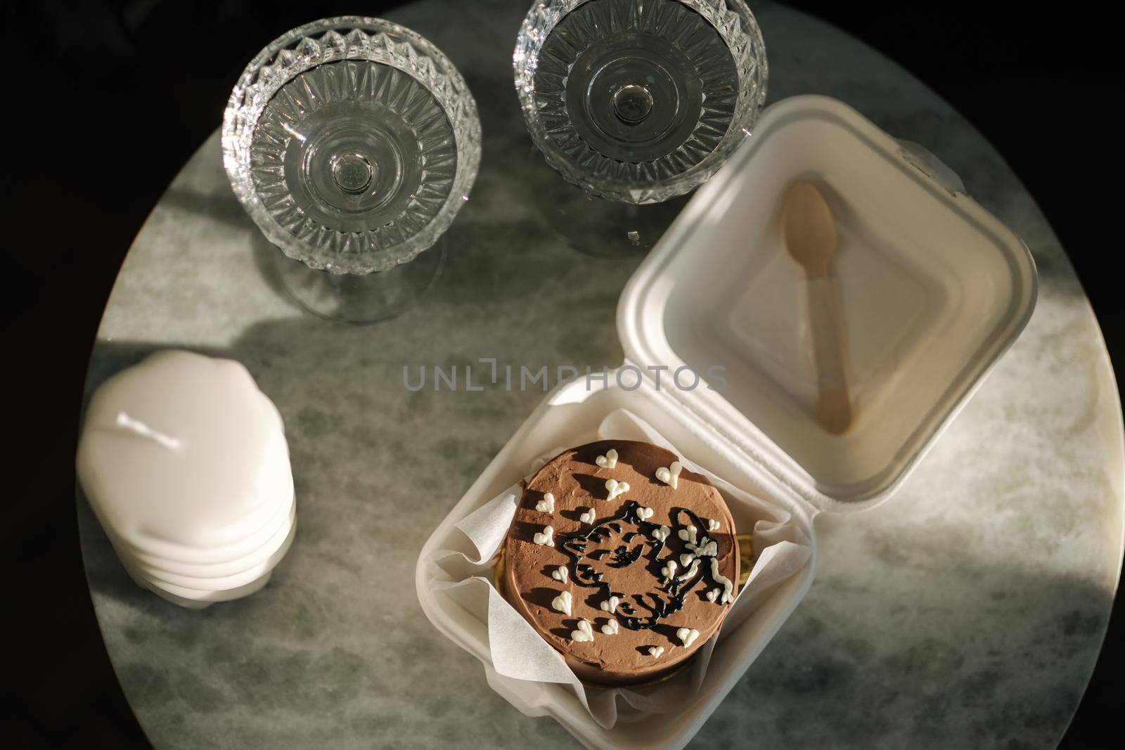 Top view of Chocolate bento cake in eco box with wooden spoon. White candle and chmpagne glasses on luzury marble table.