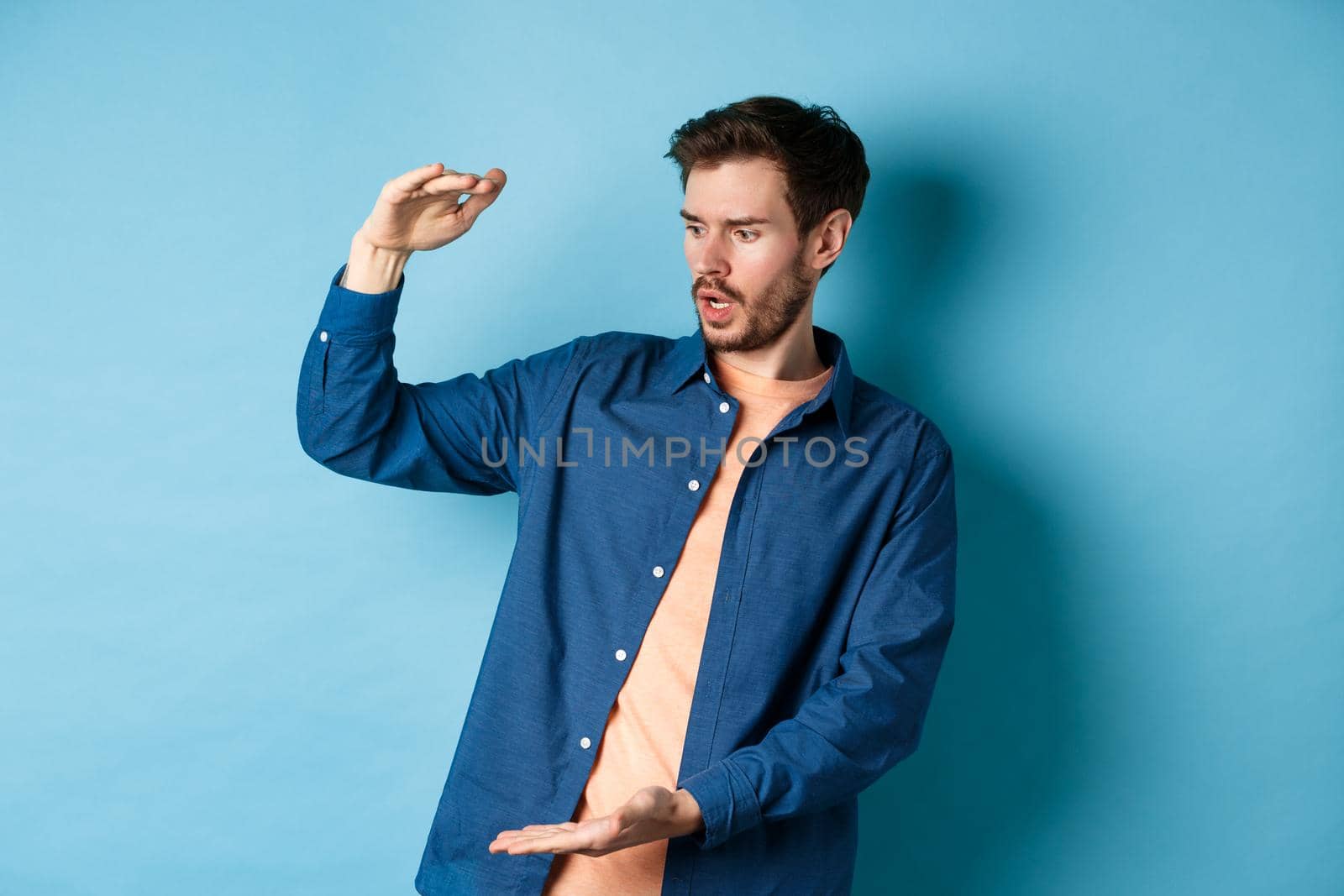 Shocked young man looking at empty space and showing big size object, standing on blue background.