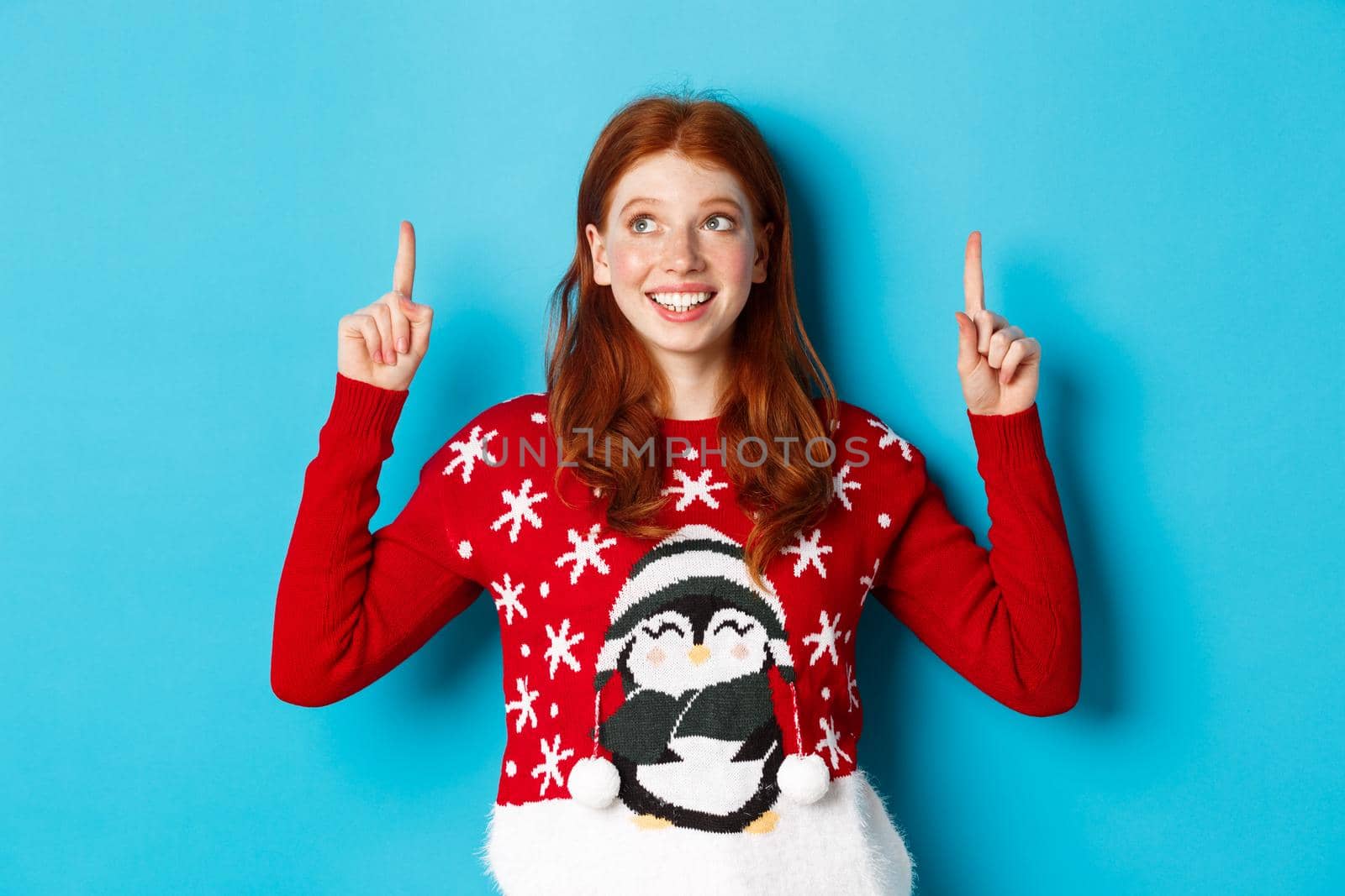 Winter holidays and celebration concept. Cheerful teen girl with red hair, looking dreamy at logo, pointing fingers up, showing advertisement, standing over blue background.