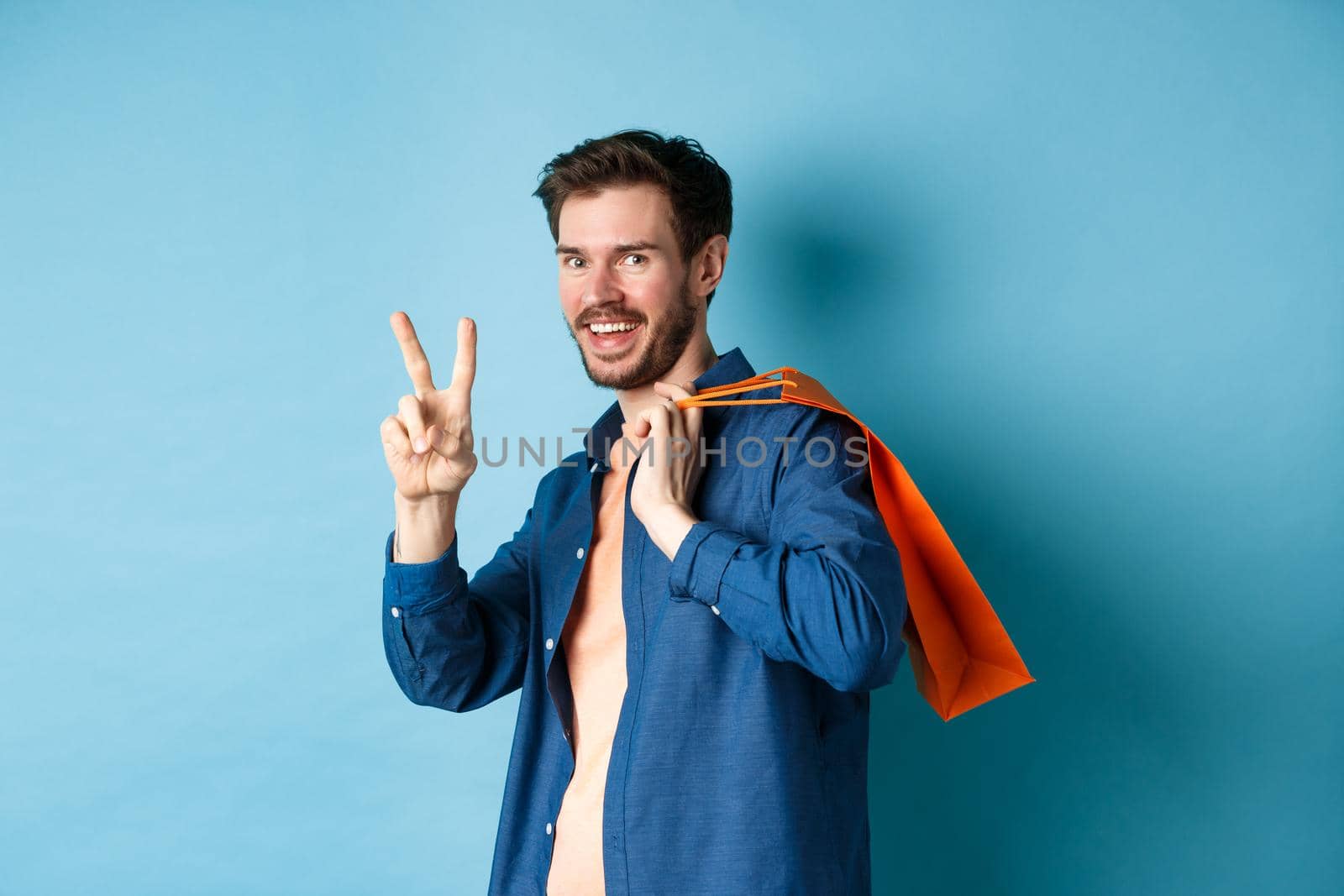 Cheerful guy holding orange shopping bag on shoulder, smiling and showing peace sign, standing on blue background. Copy space