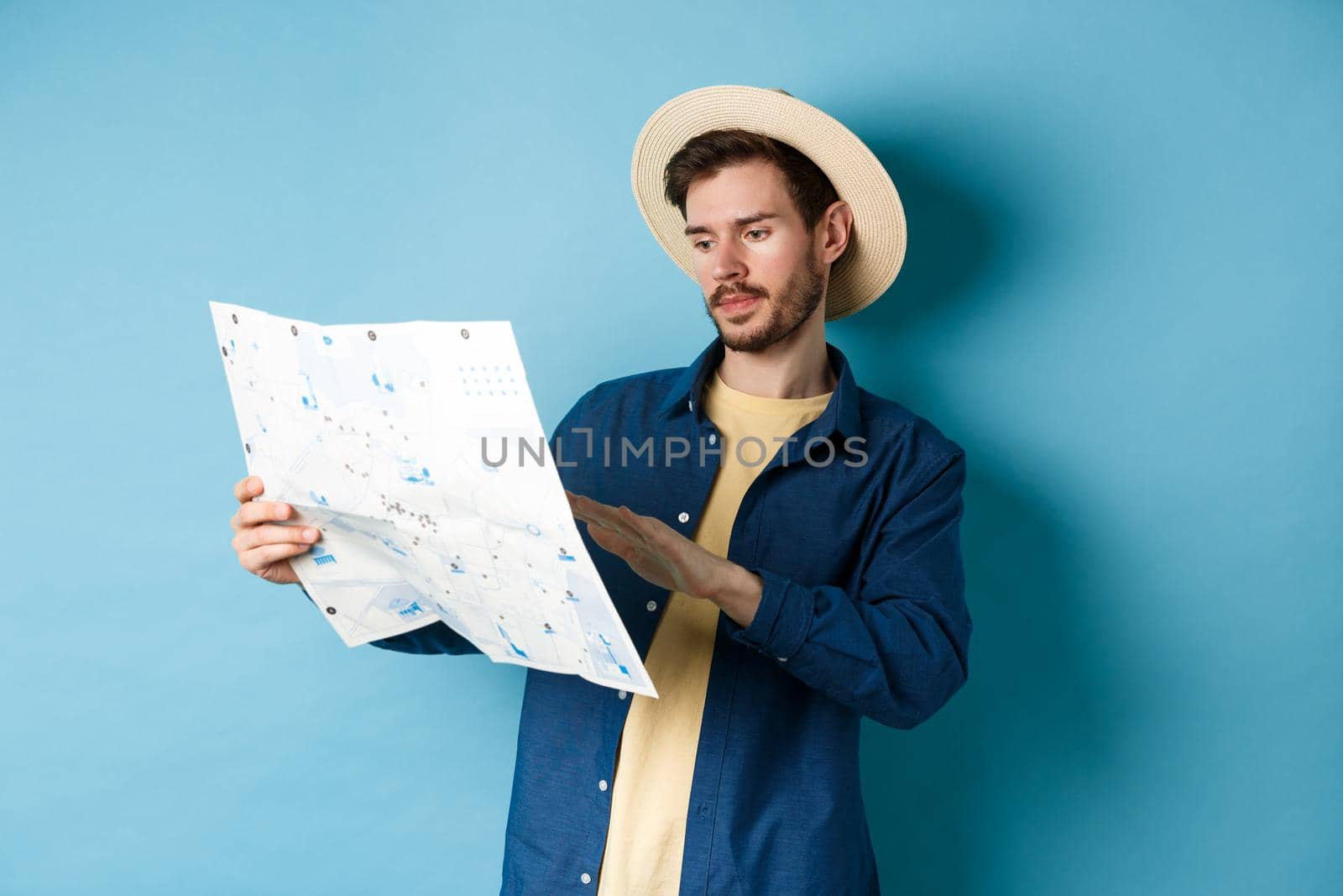 Image of handsome caucasian man looking at travel map, studying route during summer vacation, standing in straw hat on blue background.