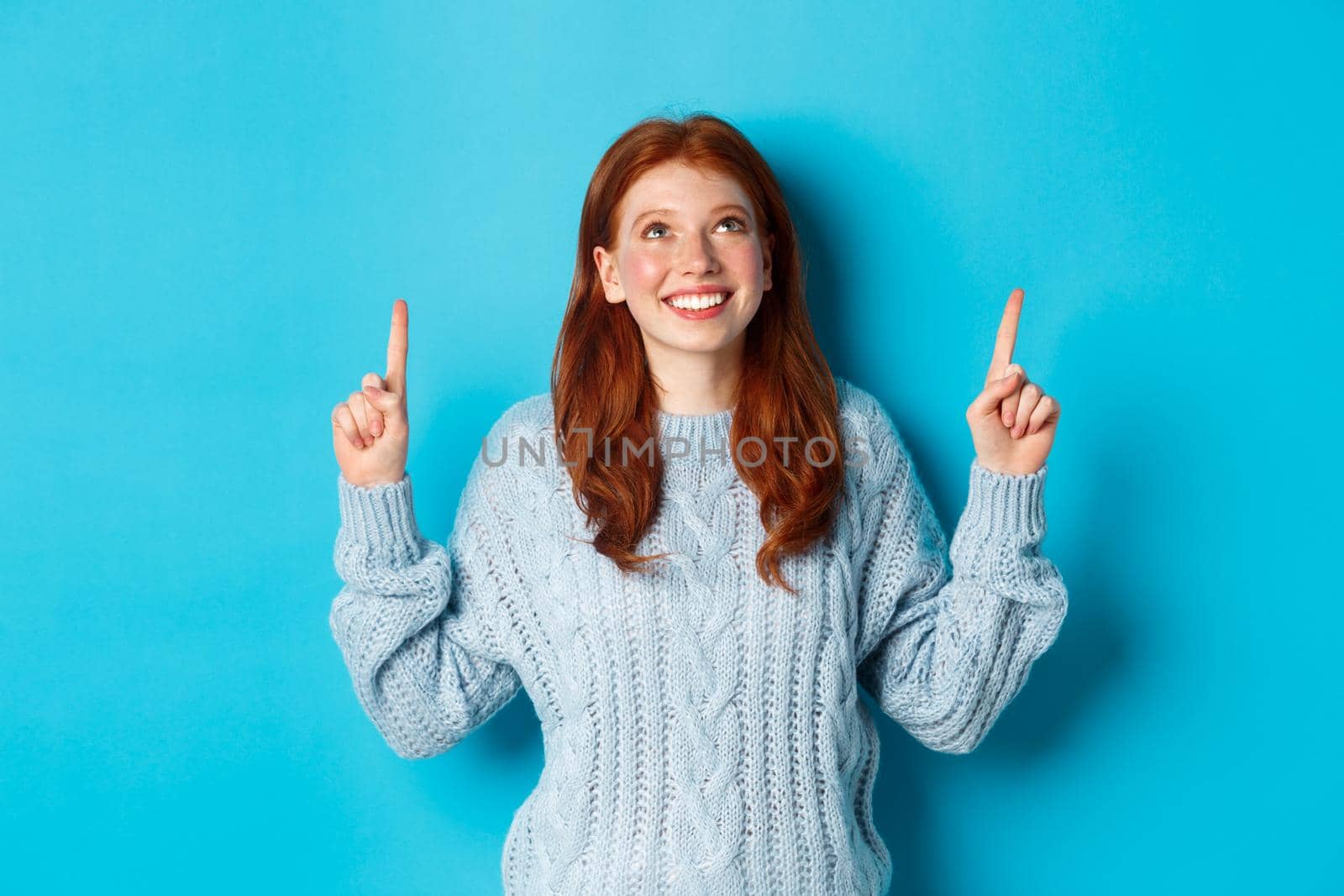 Winter holidays and people concept. Cute teenage redhead girl pointing fingers up, looking at top promo and smiling amused, standing over blue background.
