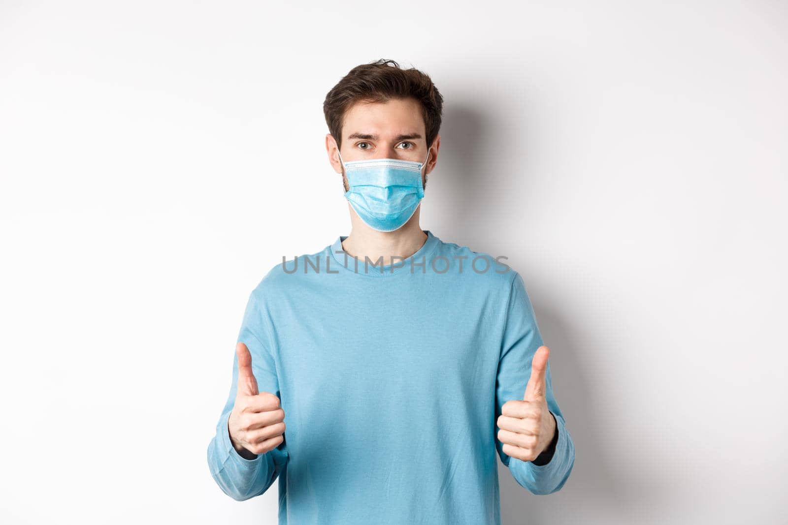 Covid-19, pandemic and social distancing concept. Young man in casual clothes and medical mask, showing thumbs up and looking serious, protecting from virus during quarantine by Benzoix