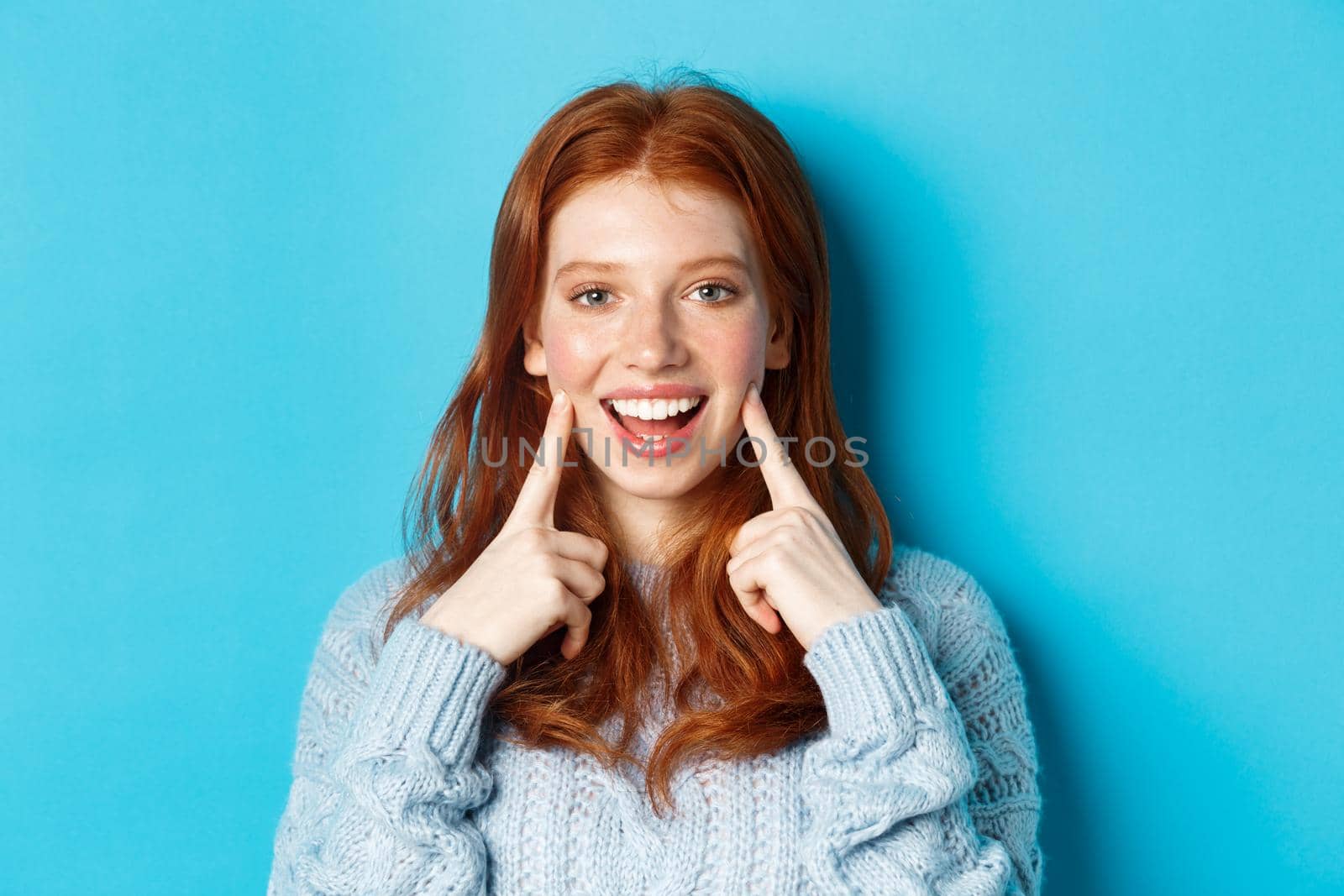 Close-up of cheerful teenage girl with red hair and freckles, poking cheeks, showing dimples and smiling with white teeth, standing over blue background by Benzoix