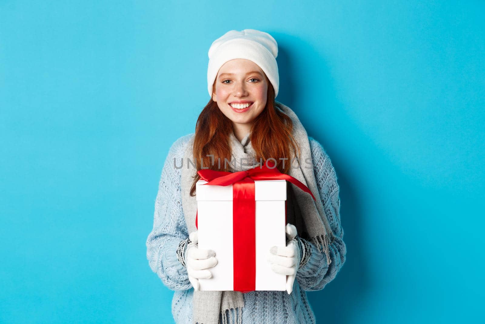 Winter holidays and Christmas sales concept. Cute redhead teeanage girl in beanie, sewater and scarf holding gift in wrapped box, smiling, wishing merry xmas, standing over blue background by Benzoix