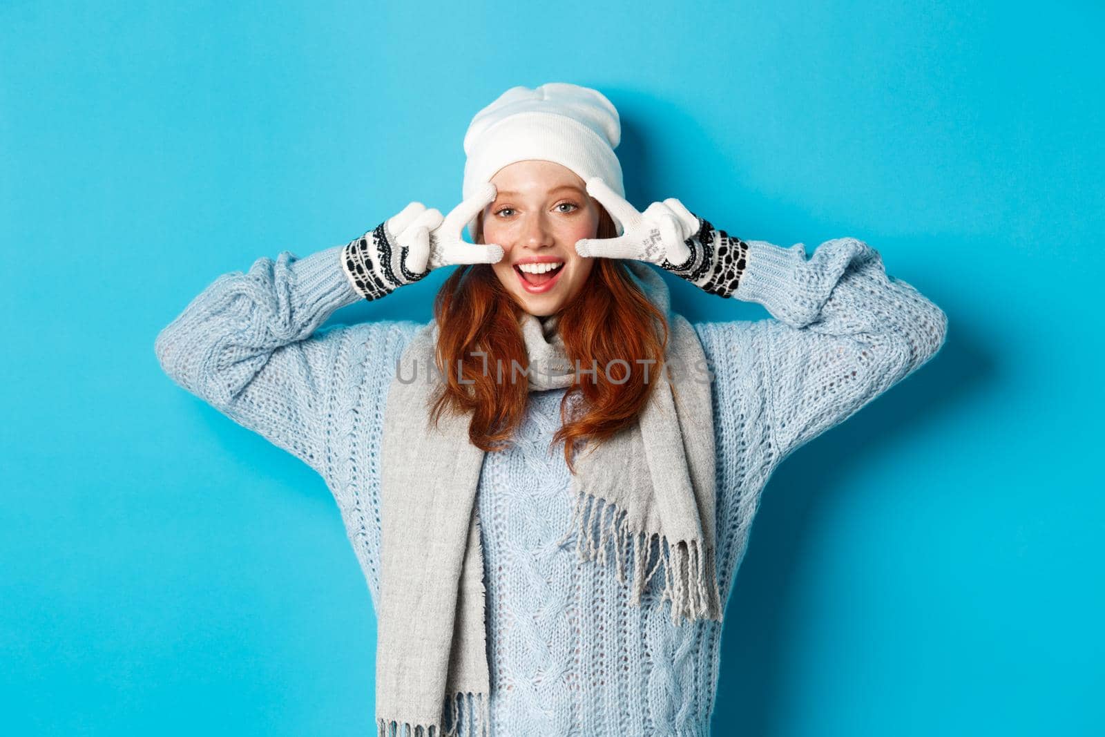Winter and holidays concept. Cute redhead teen girl in beania, gloves and sweater showing peace sign, looking left at camera and wishing merry christmas, standing against blue background.