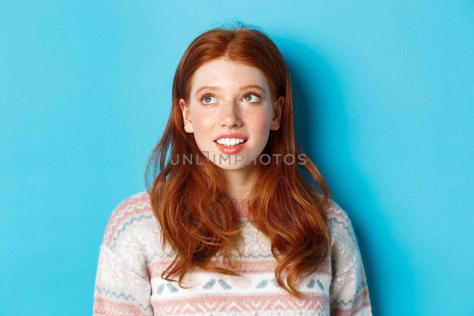 Close-up of dreamy teen girl with red hair, looking at upper left corner and smiling, standing against blue background.